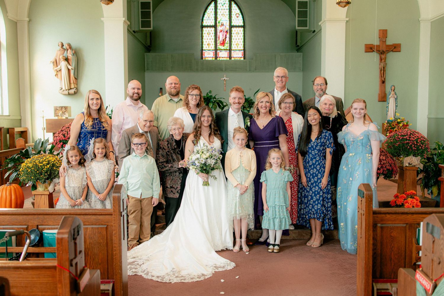 Bride and groom pose with large family group inside a charming decorated church during fall wedding ceremony celebration.