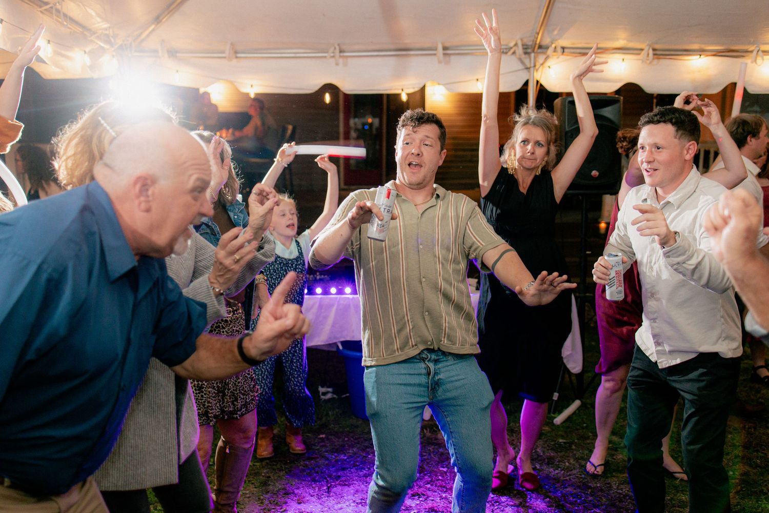 Wedding guests dance enthusiastically under a tent with purple lighting at an outdoor reception.