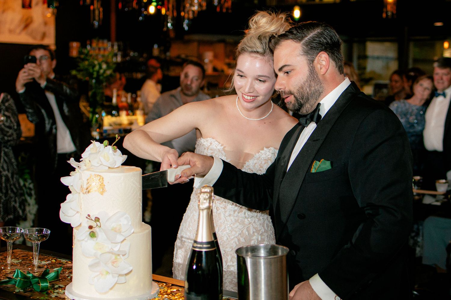 Bride and groom cutting elegant white wedding cake with champagne bottle nearby at reception.