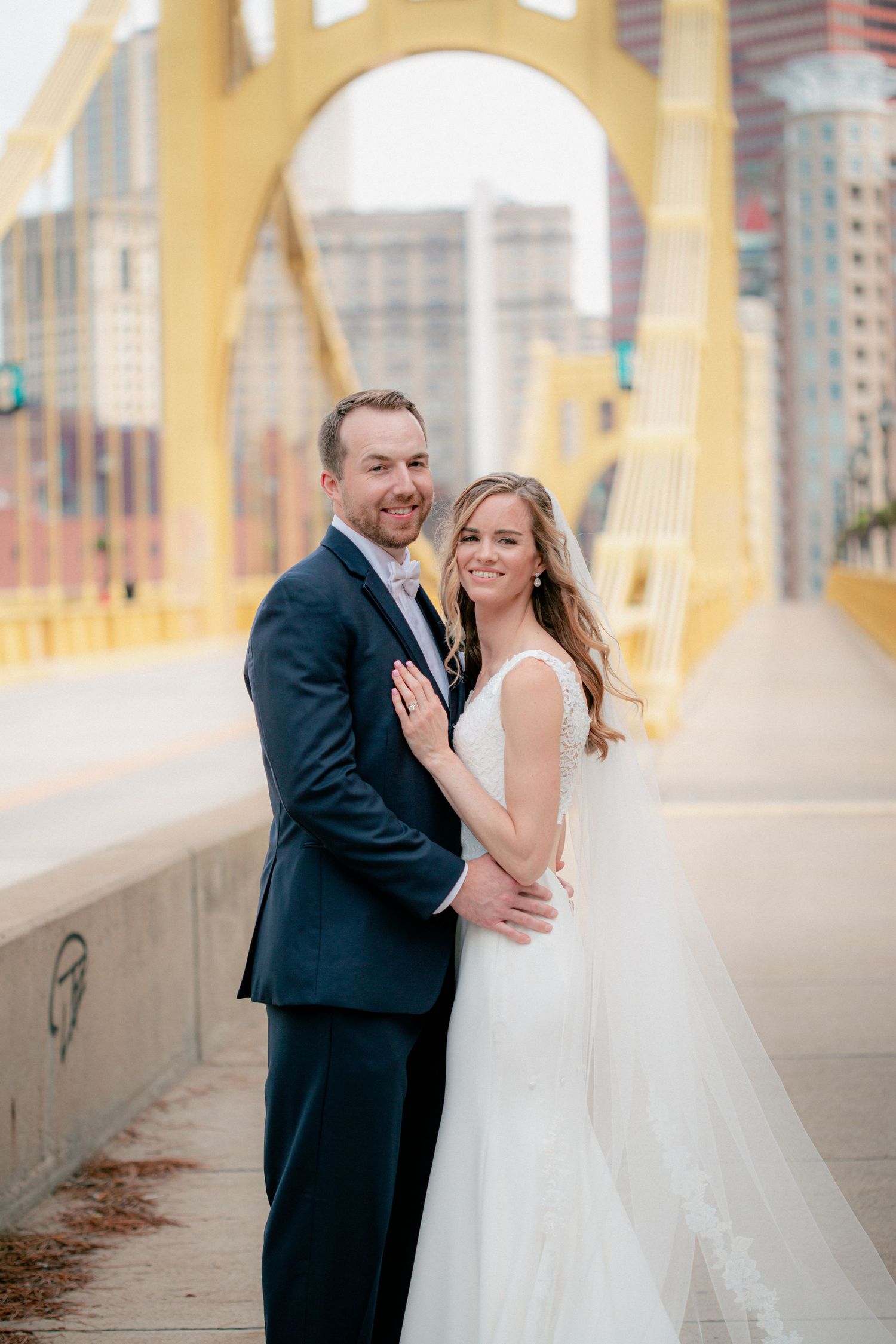 Happy newlyweds embrace and smile on Pittsburgh's yellow bridge with city skyline behind them.