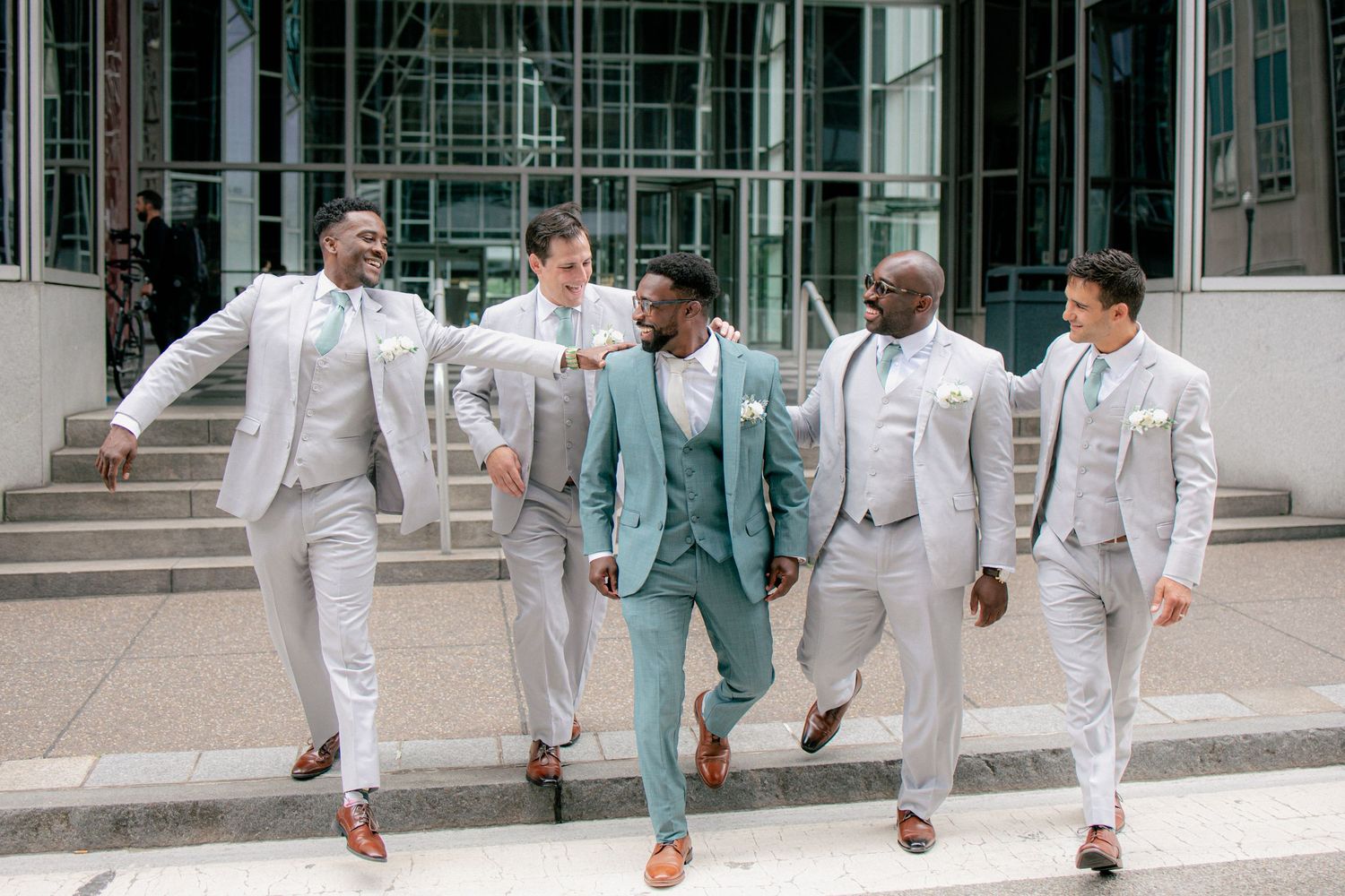 Groom in teal suit walks confidently with four groomsmen in gray suits outside modern building.