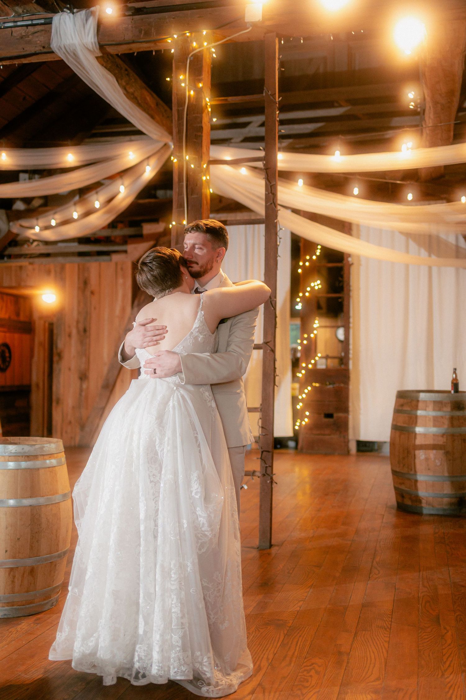 Bride and groom share first dance in rustic barn venue with fairy lights, white draping, and wooden wine barrels.