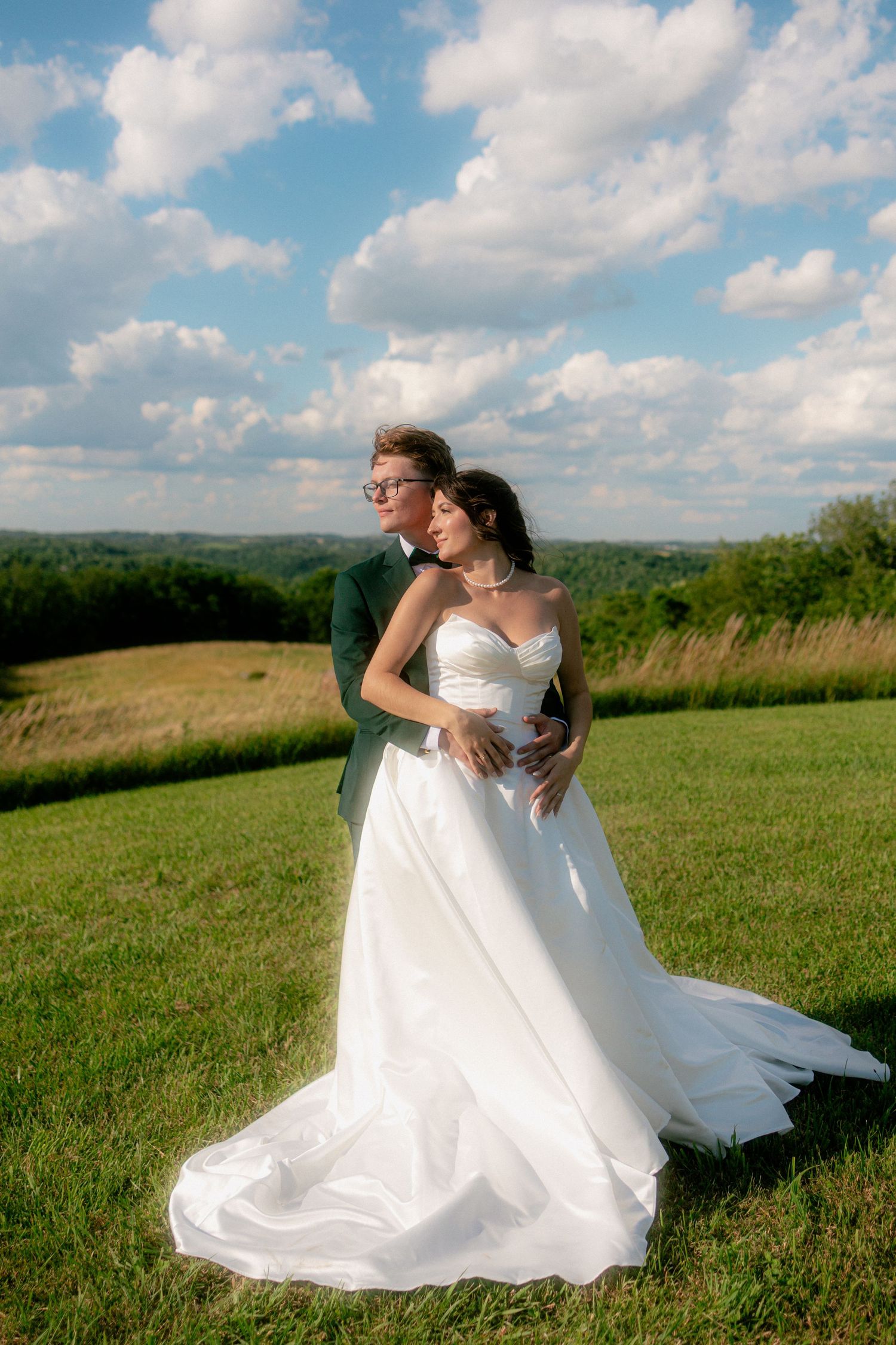 A bride in a white gown and groom in a green suit embrace in a sunny green field with rolling hills and blue sky.