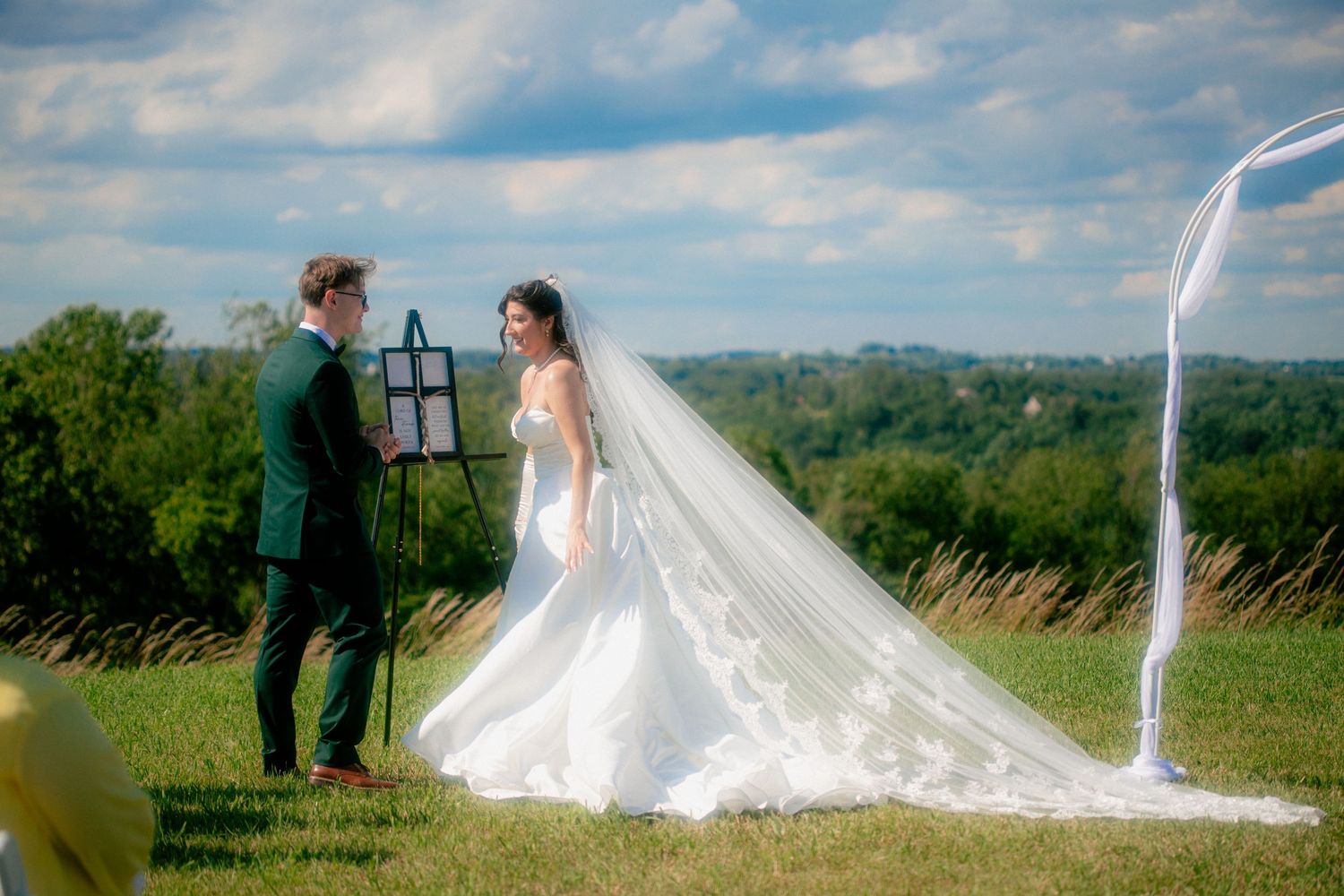 Bride in white gown with long veil stands beside groom in green suit during outdoor wedding ceremony on lush green hillside.