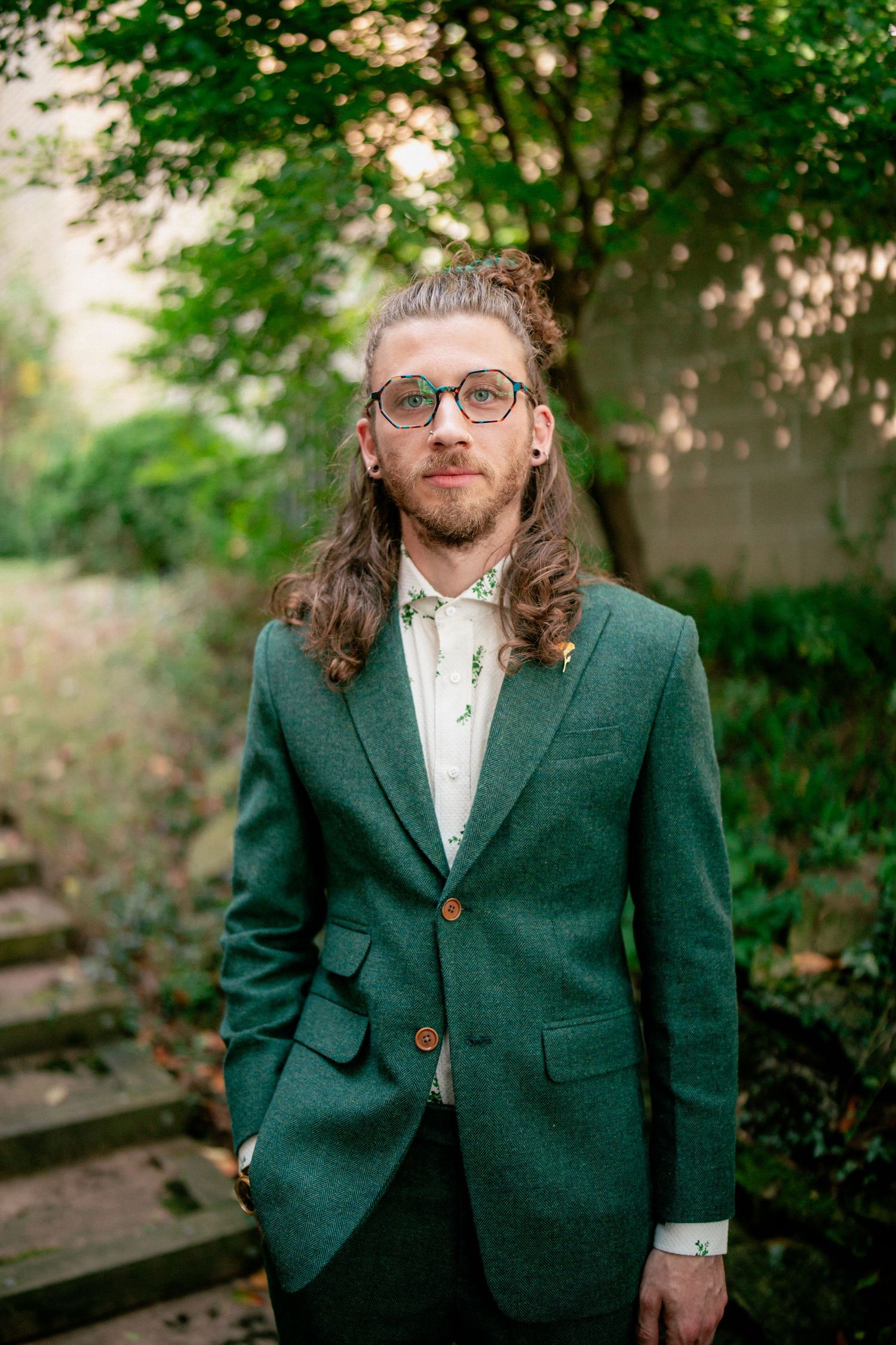 Groom with long hair and glasses wearing green tweed suit stands outdoors among lush green trees.