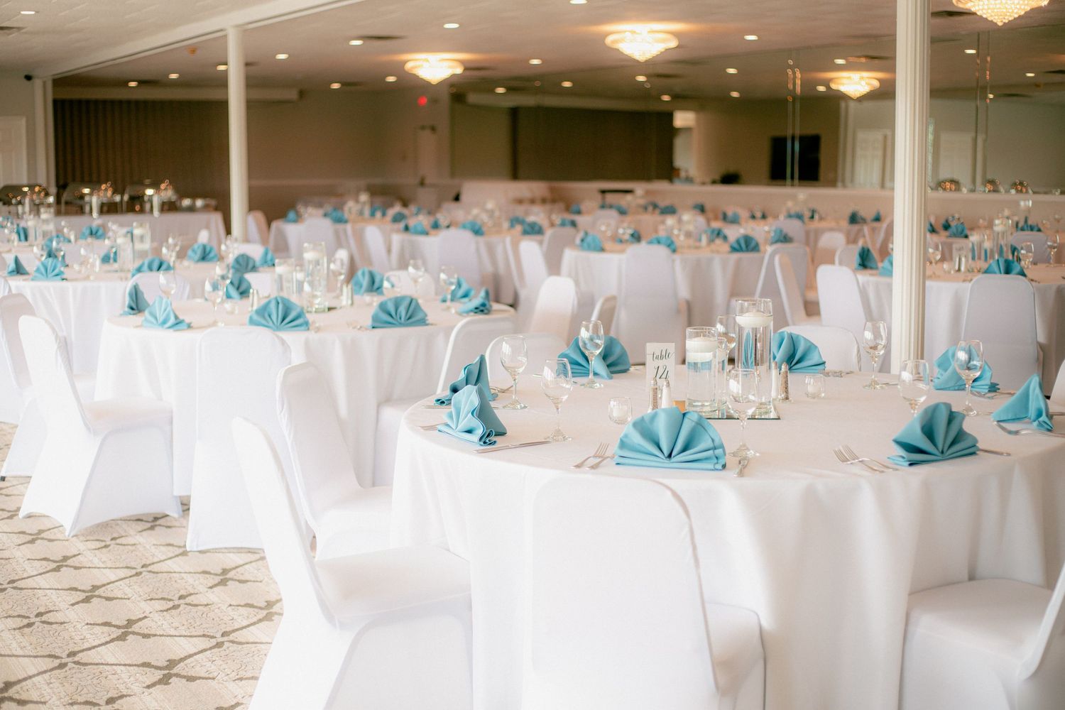 Elegant wedding reception hall with round tables, white chair covers, and turquoise folded napkins.