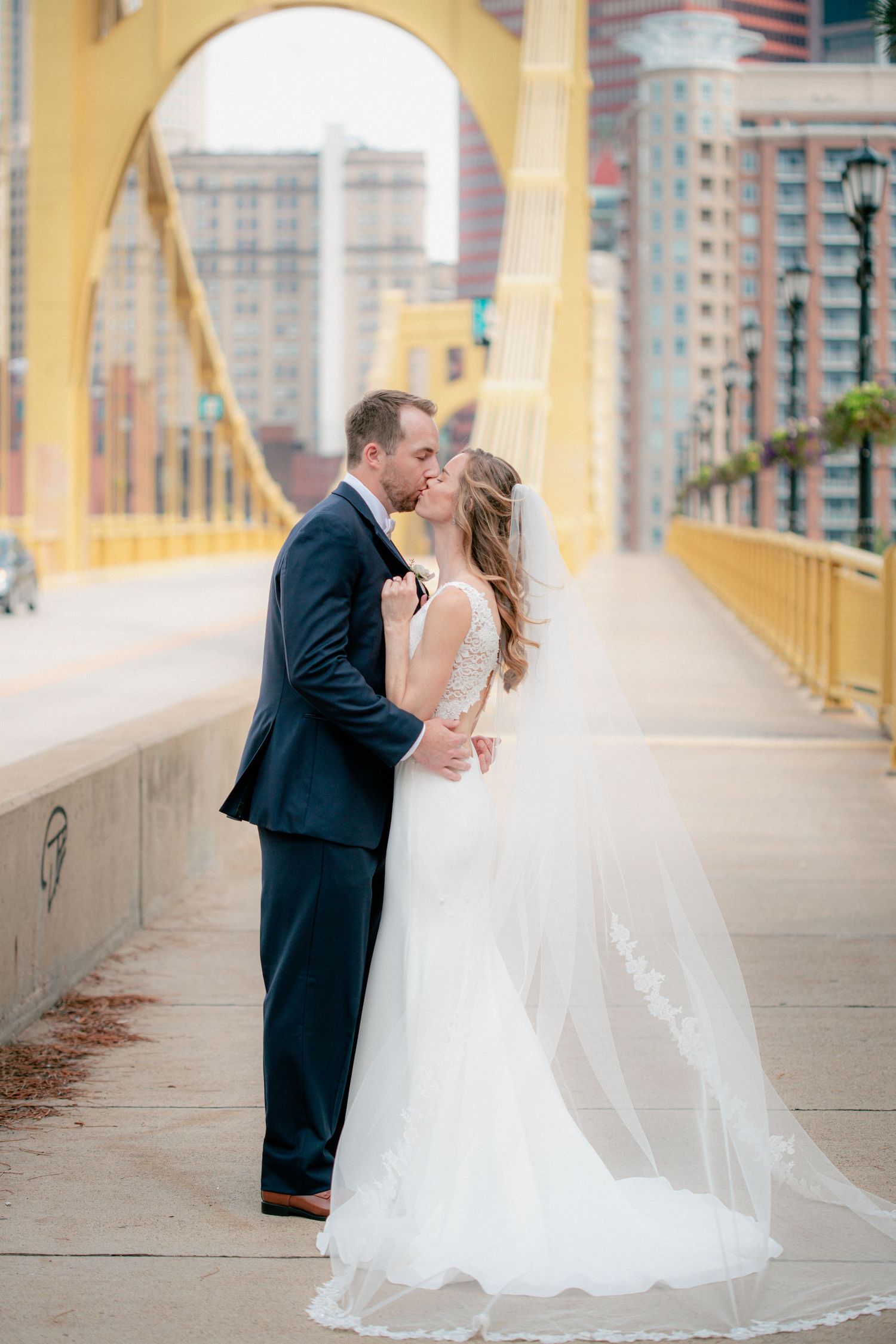 Groom kisses bride on a yellow Pittsburgh bridge, her lace cathedral veil trailing behind.