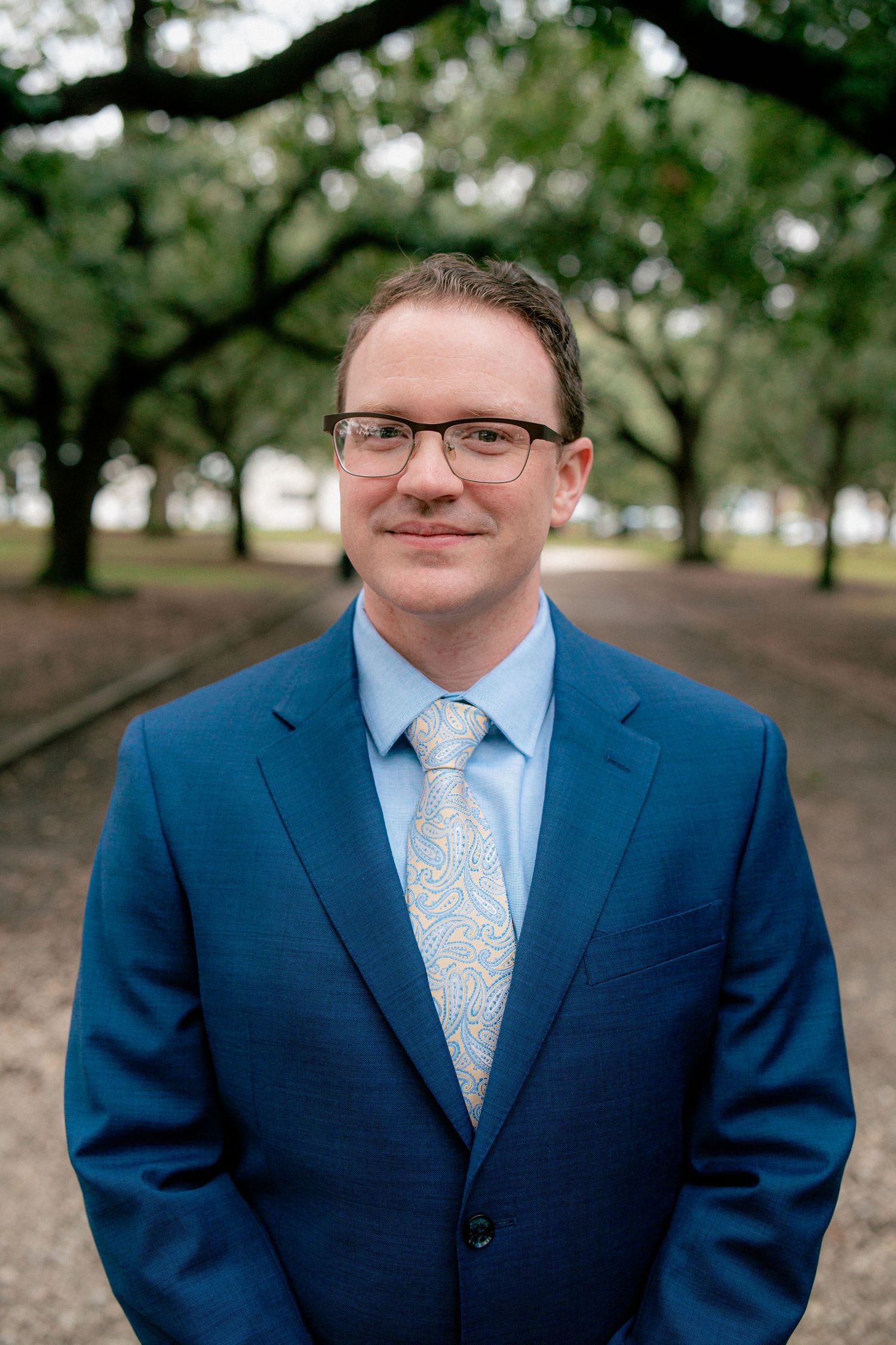 Portrait of a man in a blue suit and paisley tie wearing glasses, standing in a tree-lined park.
