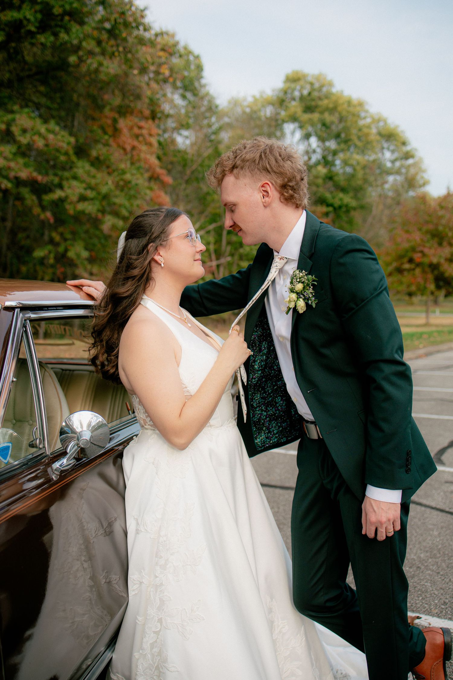 Bride playfully pulls groom's tie beside a classic vintage car in an autumn outdoor wedding portrait.