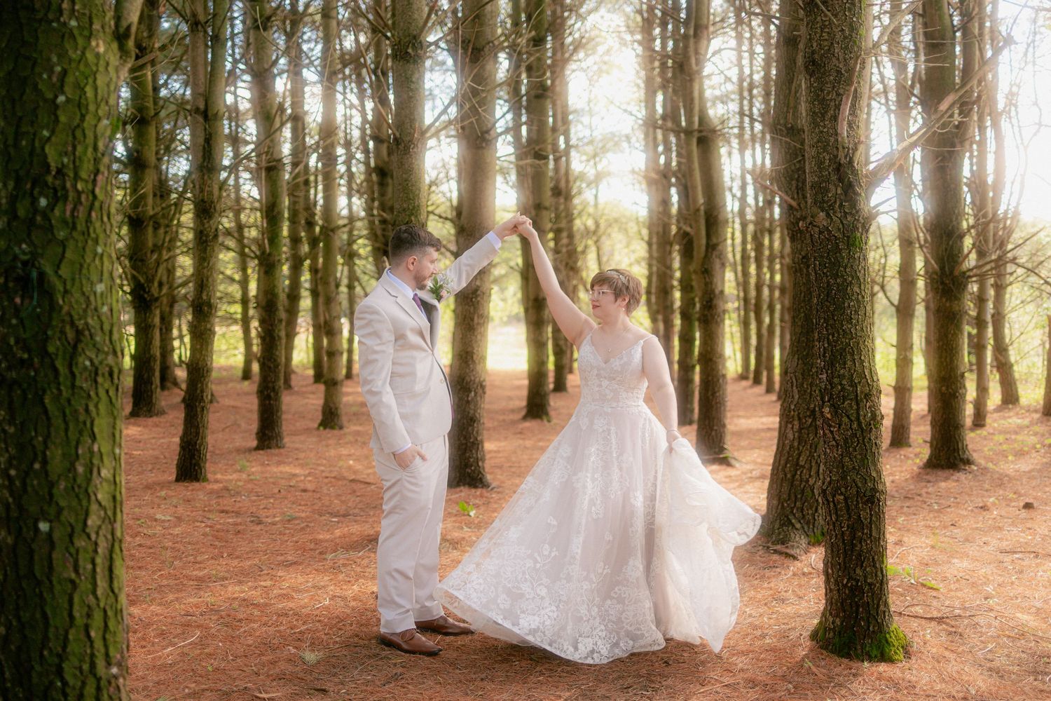 Bride and groom dancing among tall trees in sunlit forest, bride in flowing white gown, groom in light suit.