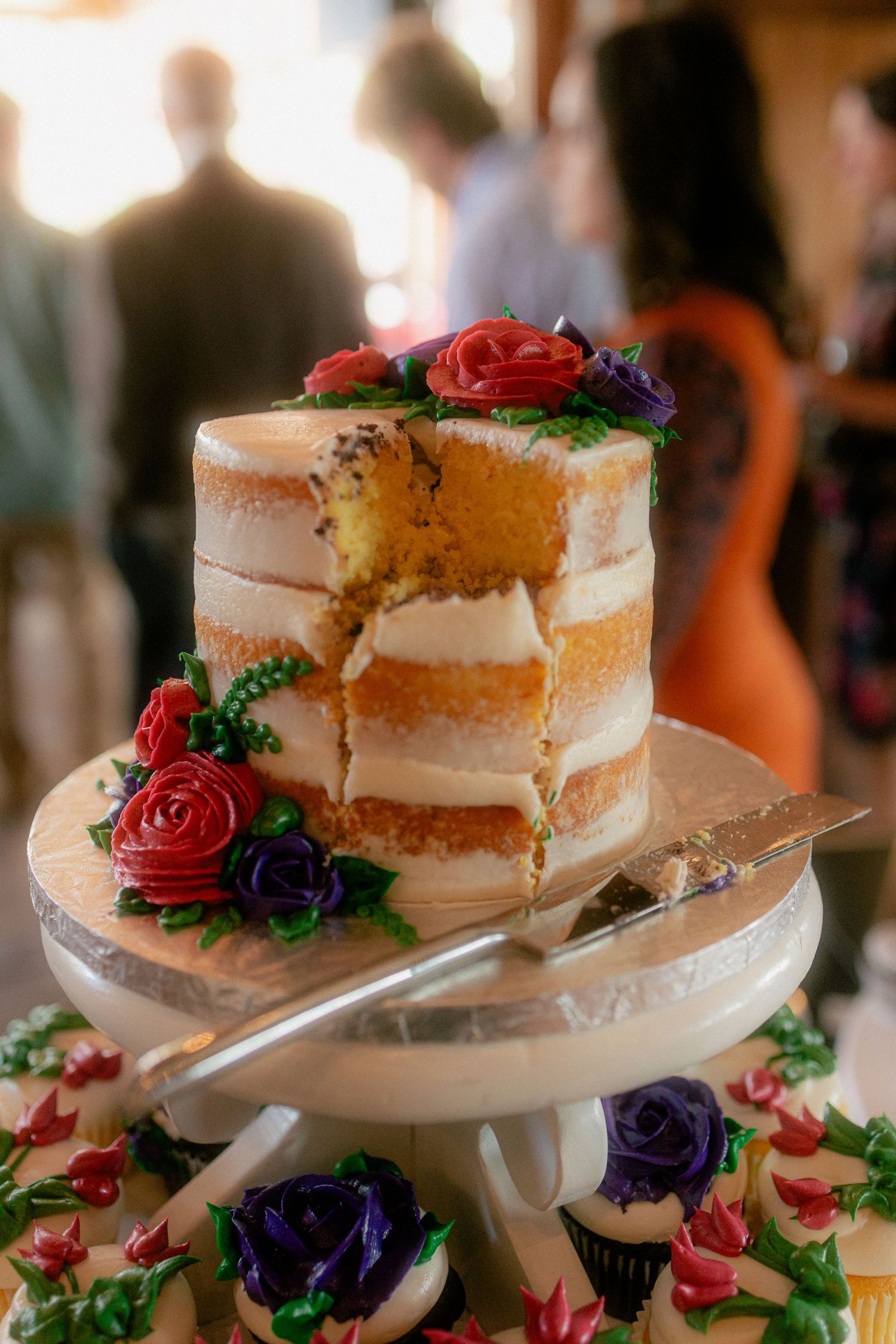 Elegant naked wedding cake with red and purple rose decorations, partially sliced, displayed on a white cake stand.