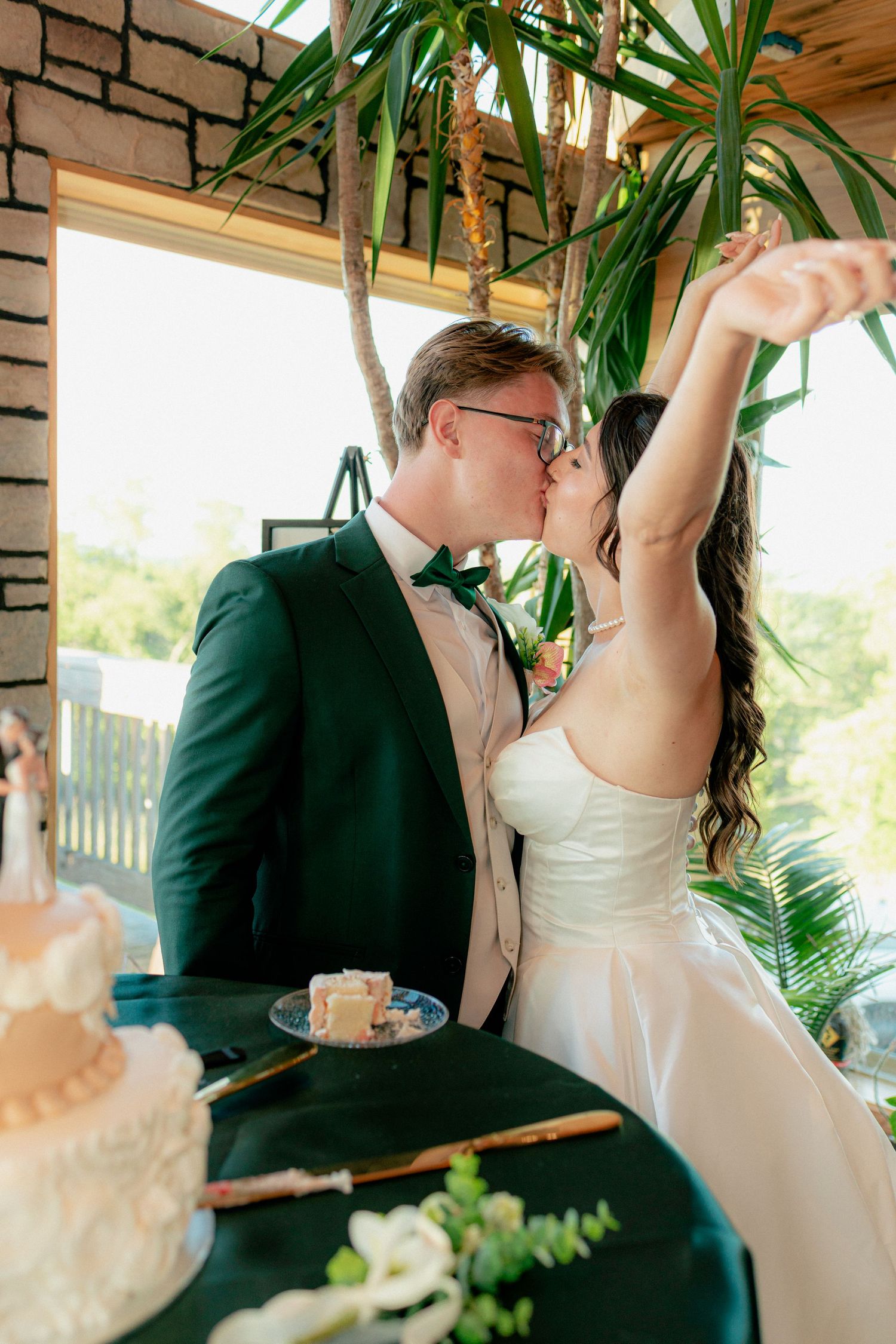 Bride and groom share a kiss at wedding cake table, bride raises arm in celebration, lush tropical greenery backdrop.