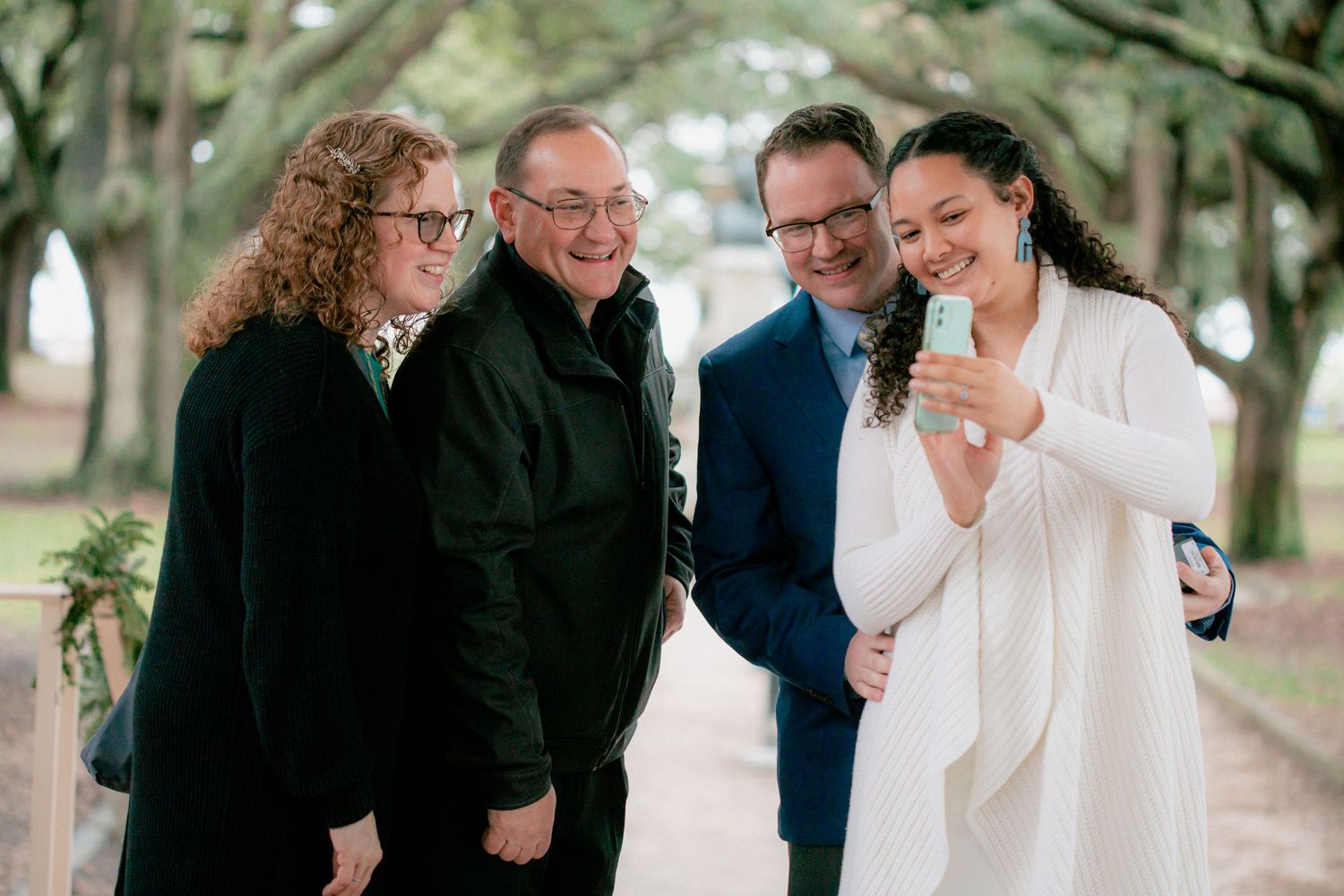Newlywed couple shares phone photos with two guests under oak trees after intimate outdoor wedding ceremony.
