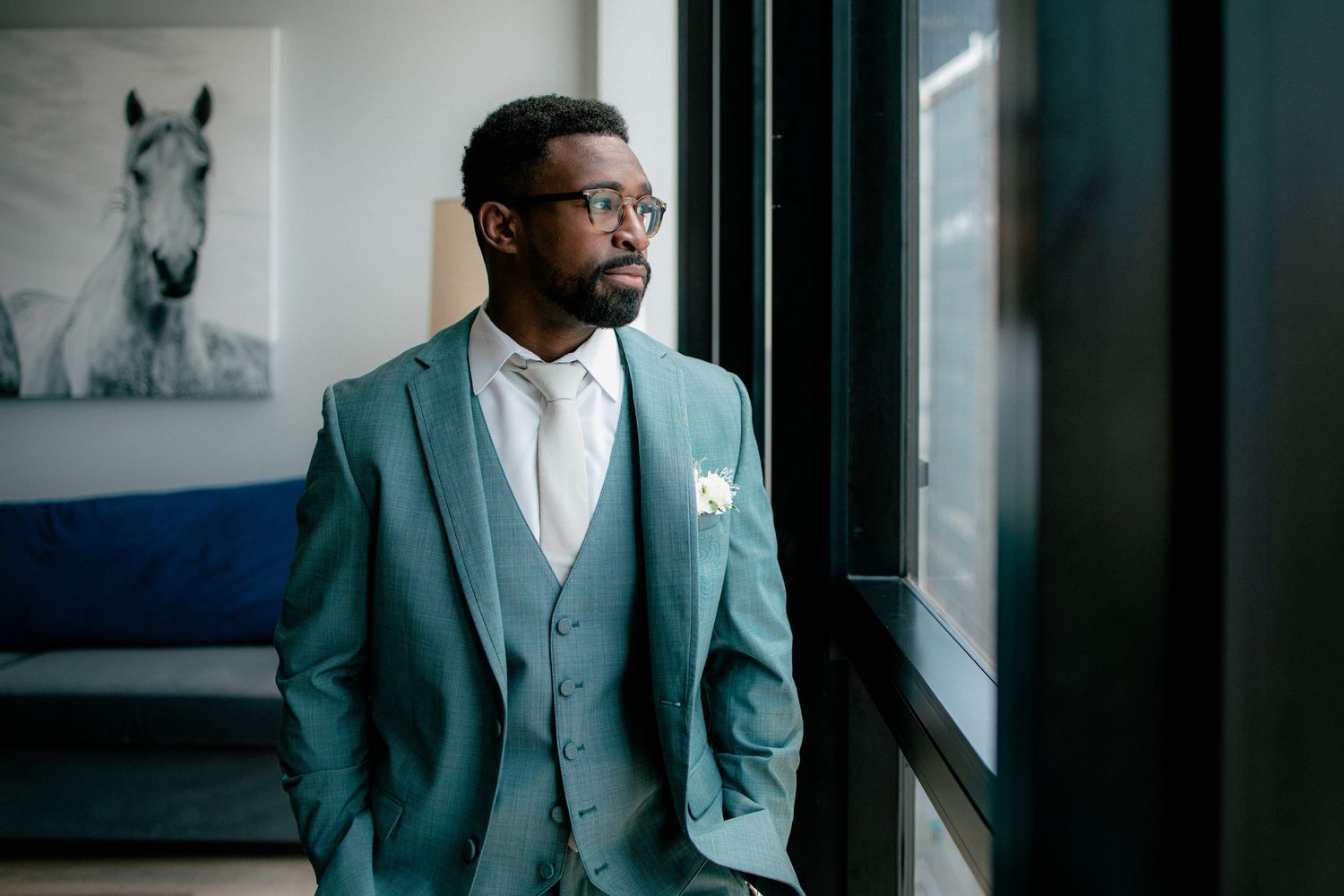 Dapper groom in teal suit and glasses gazing thoughtfully out large window.