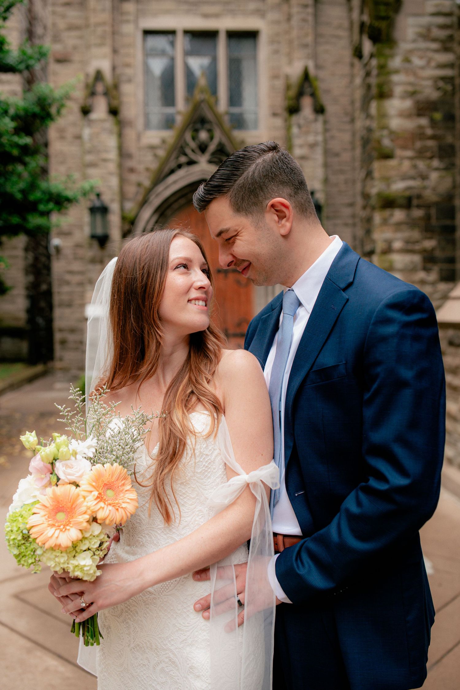 Bride holding peach floral bouquet smiles at groom in navy suit outside stone church wedding venue.