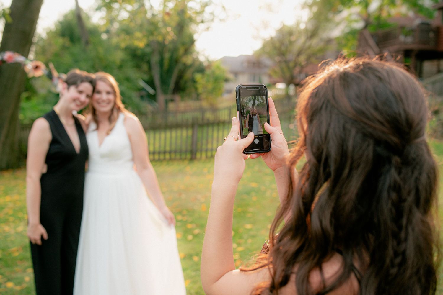 Guest photographs bride posing with woman in black dress at outdoor wedding reception.