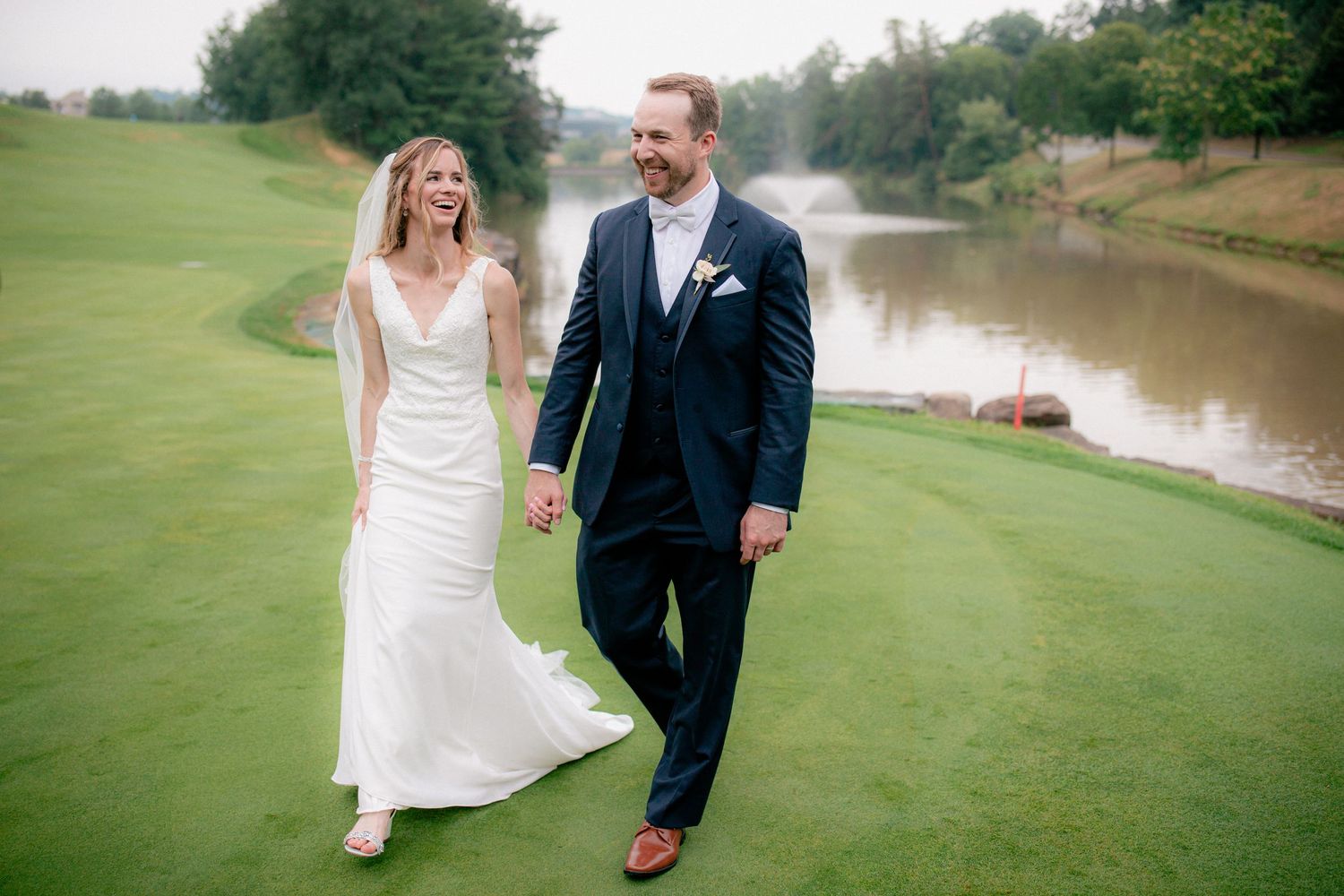 Smiling bride and groom walking hand in hand along lush golf course with river in background.