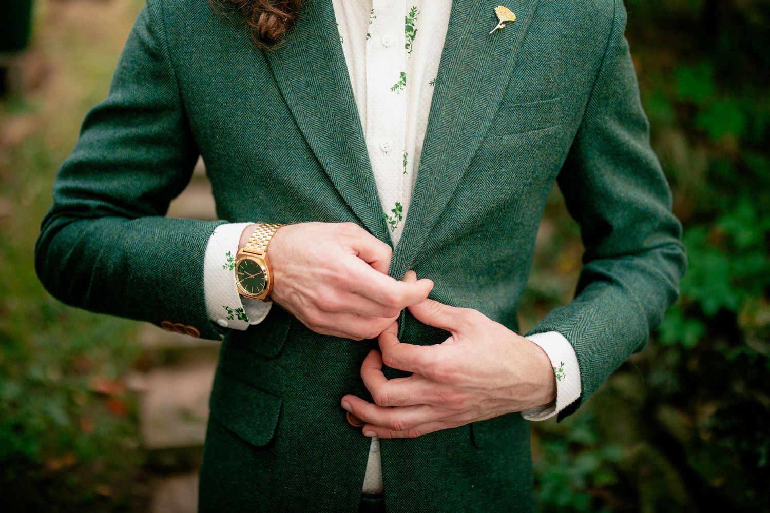Groom in green tweed suit adjusts jacket button, wearing gold watch and floral boutonniere.