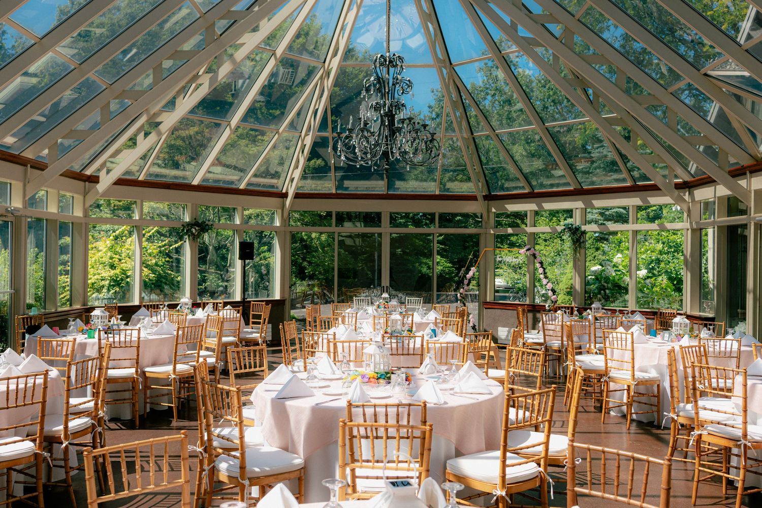 Bright conservatory wedding venue with glass ceiling, round tables, gold chiavari chairs, and garden views.