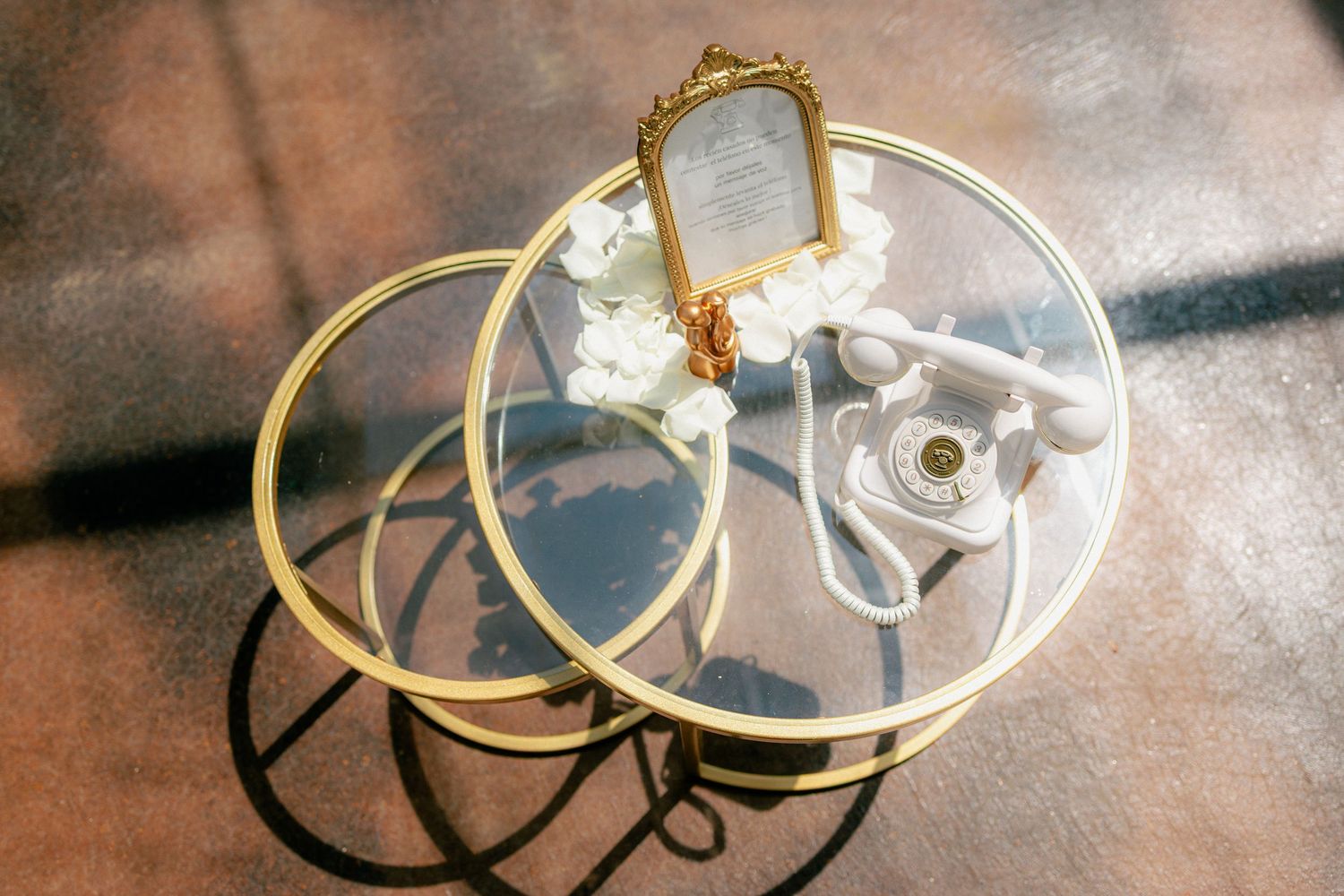 Gold glass side table with vintage white telephone, ornate mirror, and white floral arrangement.