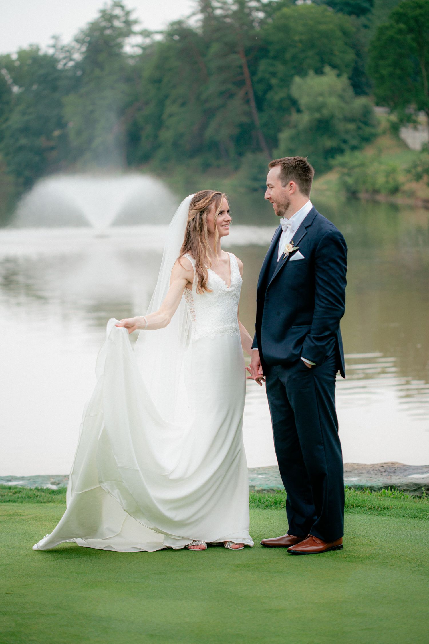 Bride and groom holding hands on golf course green with misty fountain and lake in background.