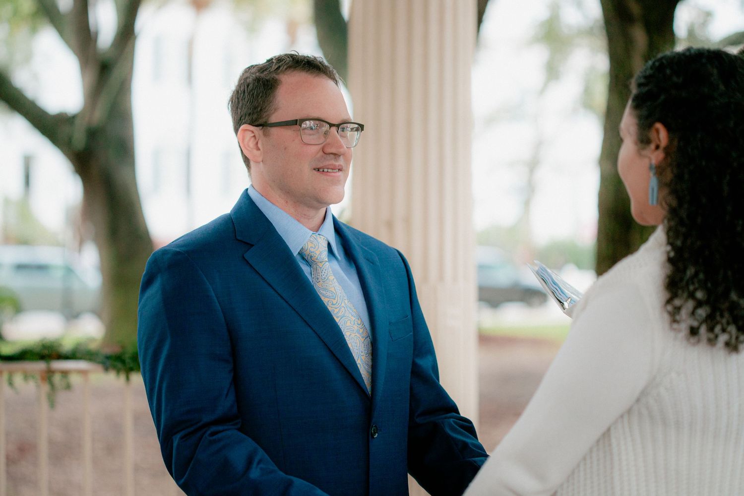 Groom in blue suit smiles warmly at bride during intimate outdoor wedding ceremony beside white columns.