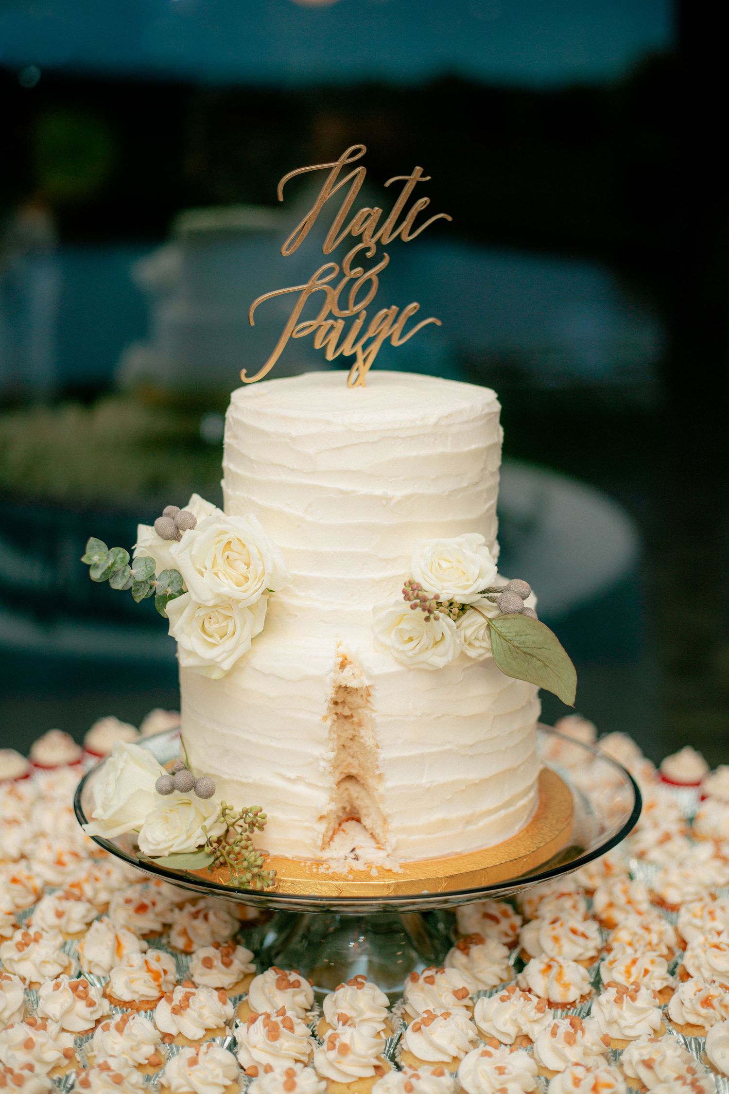 Two-tier white textured wedding cake with gold topper reading Nate and Paige, adorned with white roses.