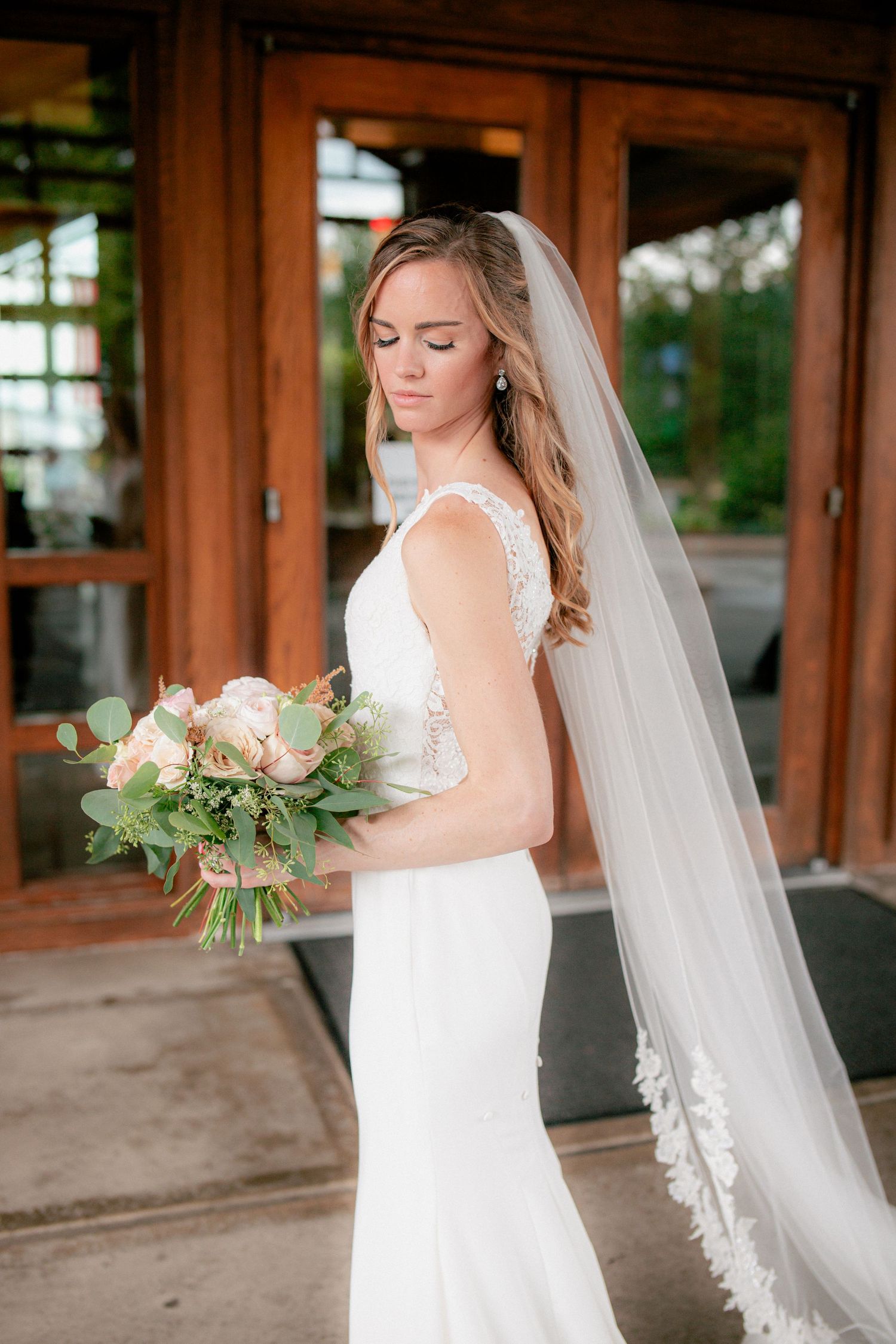 Bride in fitted lace back wedding gown with cathedral veil holding blush floral bouquet by wooden doors.