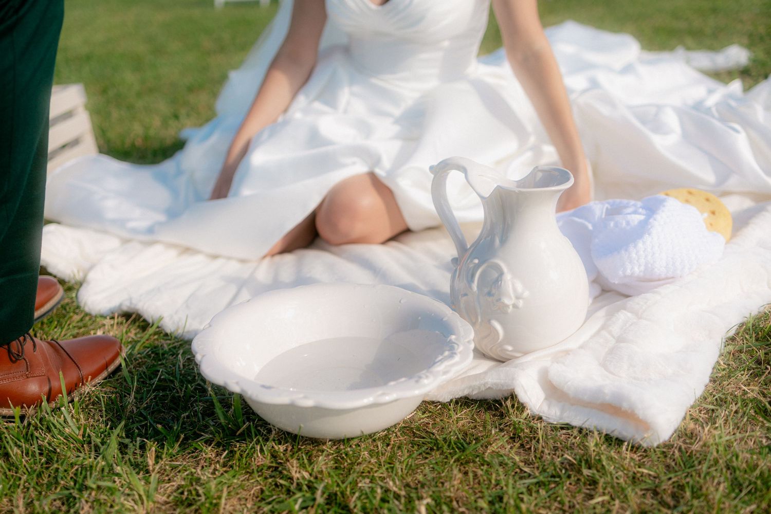 Bride in white wedding dress sitting on blanket in grass with white ceramic pitcher and basin.