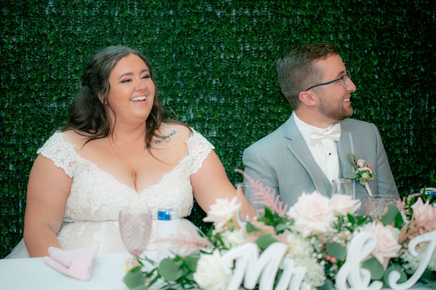 Bride and groom smile joyfully at sweetheart table adorned with pink florals and greenery backdrop.