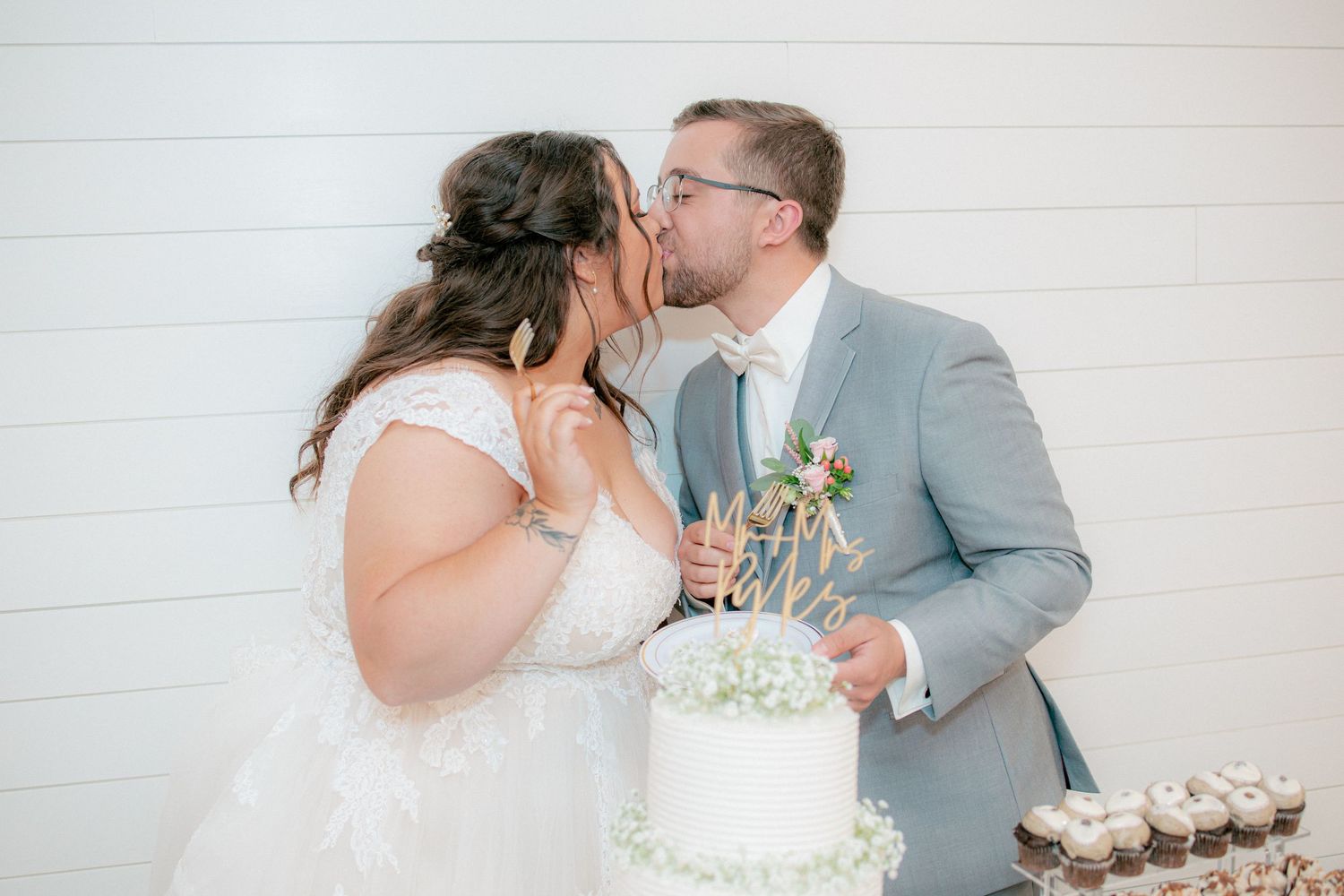 Newlyweds share a kiss beside their white wedding cake during romantic reception celebration.