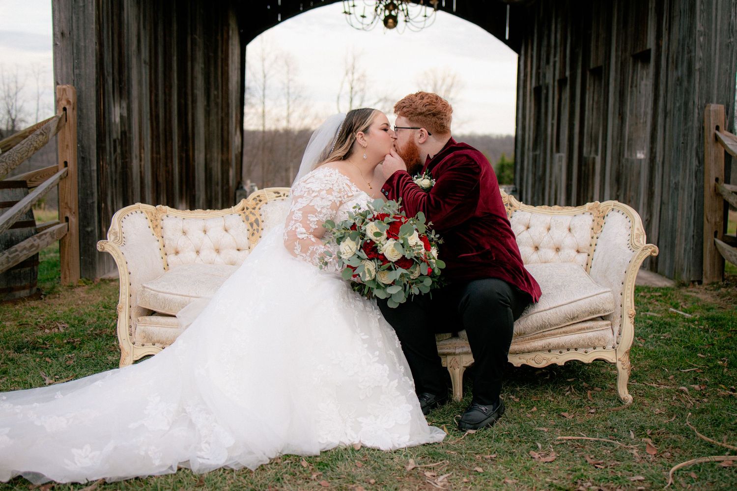 Bride and groom share a romantic kiss on a vintage ivory sofa outside a rustic barn wedding venue.