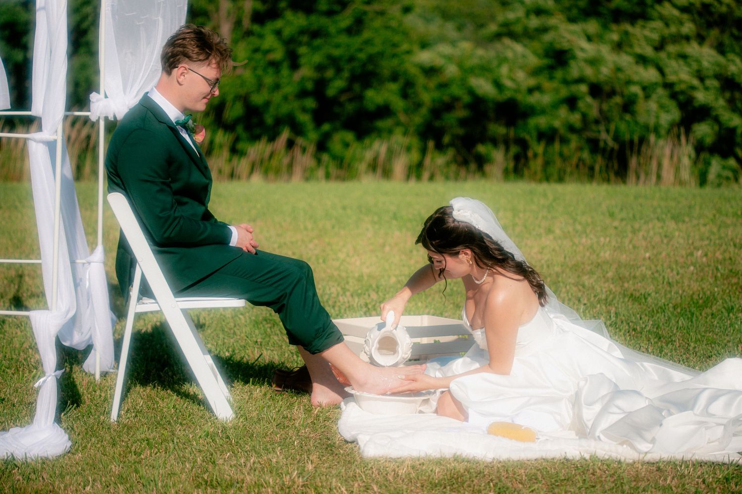Bride washing groom's feet in outdoor wedding ceremony on green grass, wearing white gown and veil.
