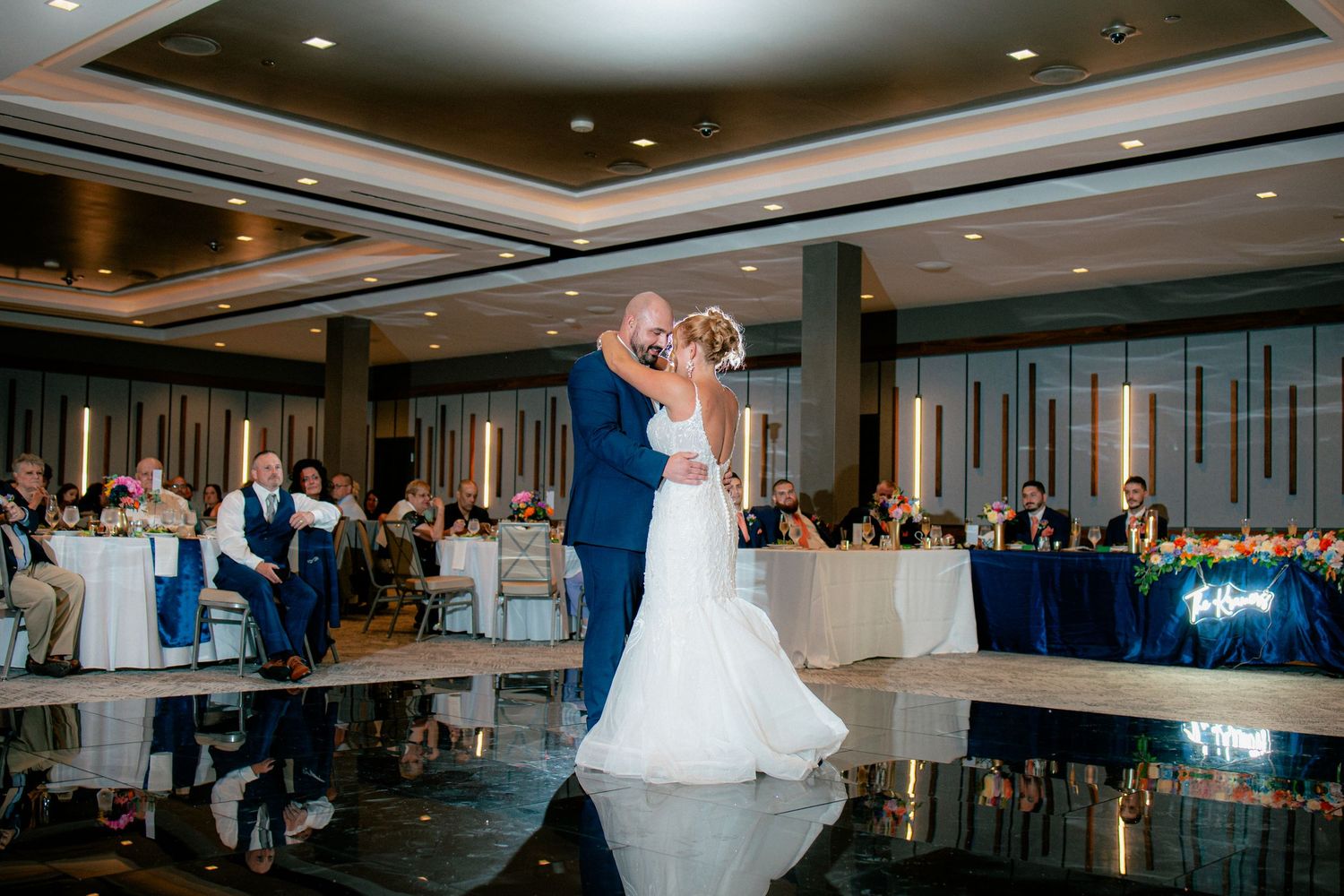 Bride and groom share romantic first dance kiss on reflective ballroom floor before seated guests.