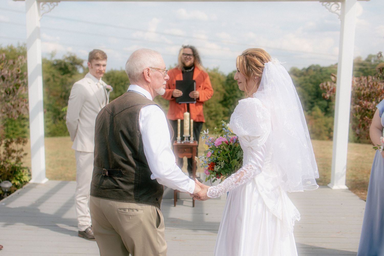 Bride and father hold hands at outdoor gazebo wedding ceremony with officiant in red standing behind.