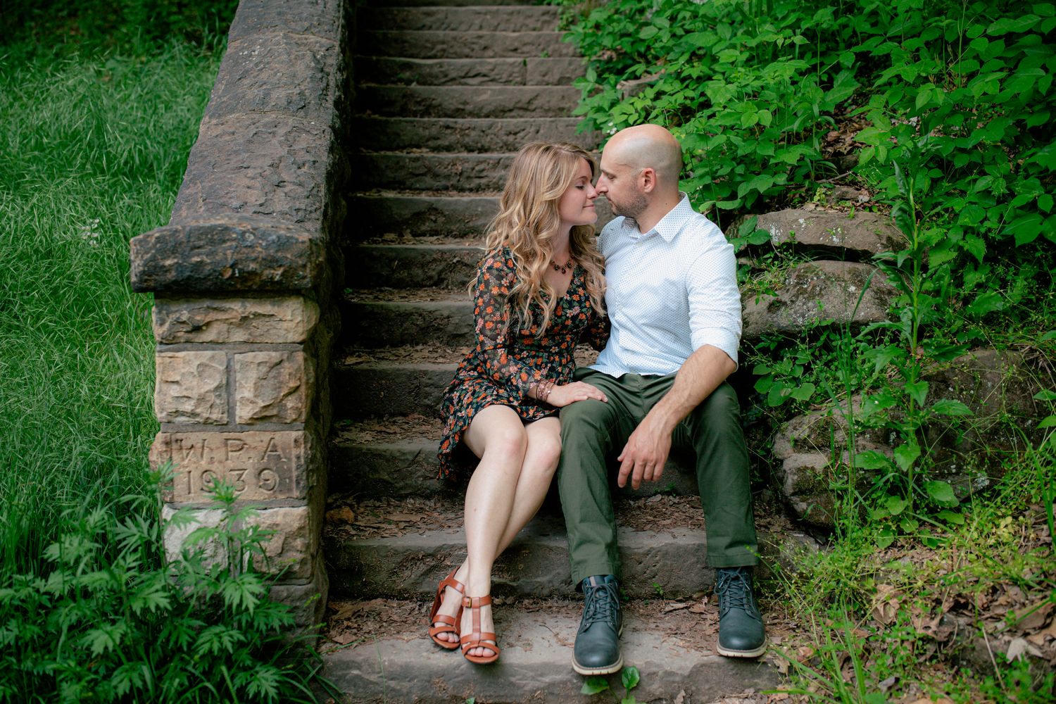 Couple sitting close together on stone steps in a lush green park, sharing a tender kiss.