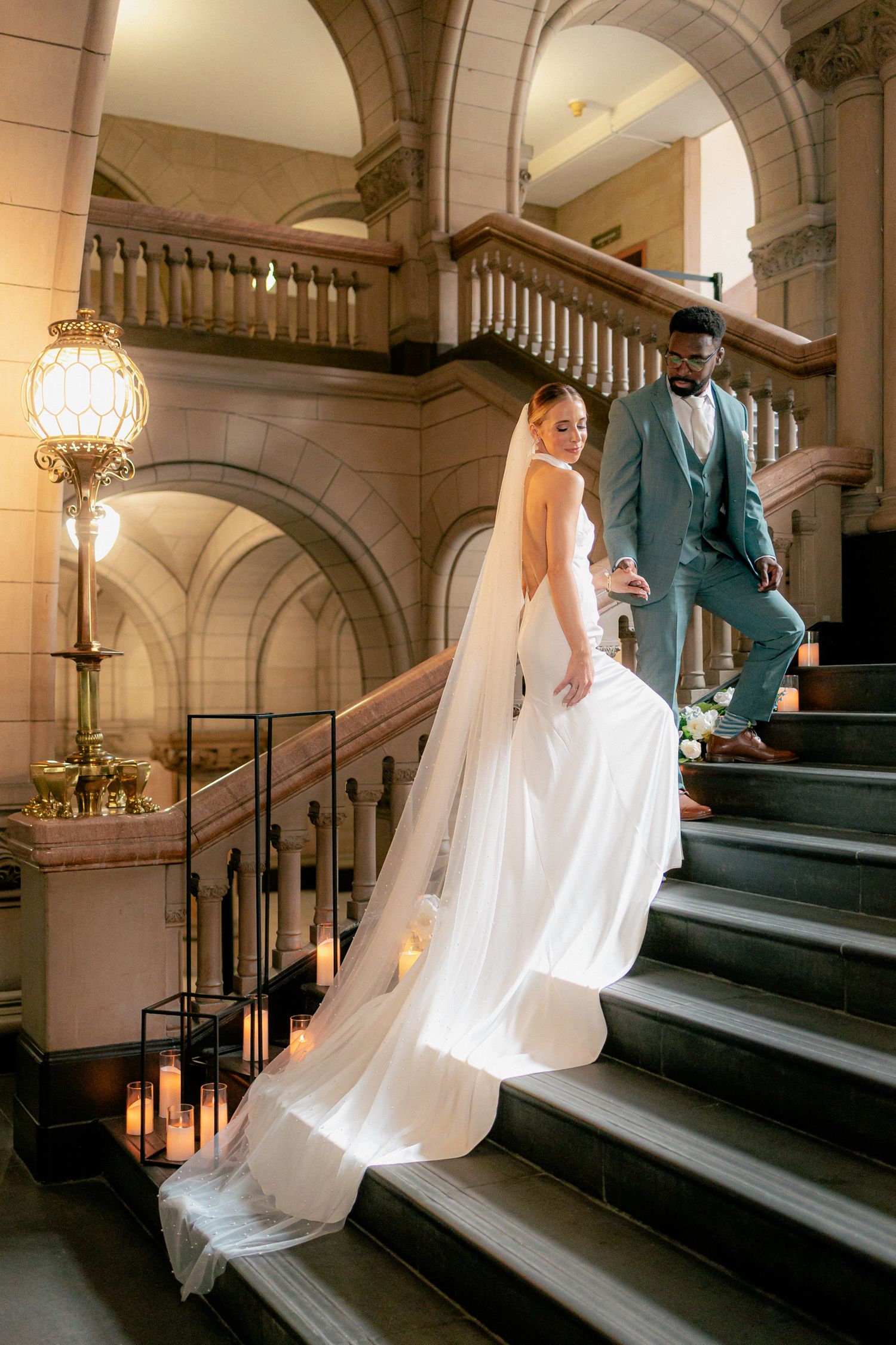 Bride in backless white gown with cathedral veil descends grand staircase with groom in teal suit.