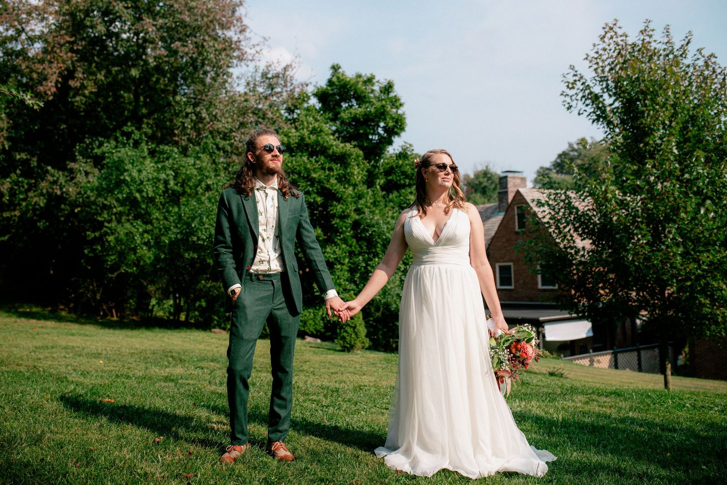 Bride and groom hold hands wearing sunglasses on sunny green lawn with rustic building behind them.