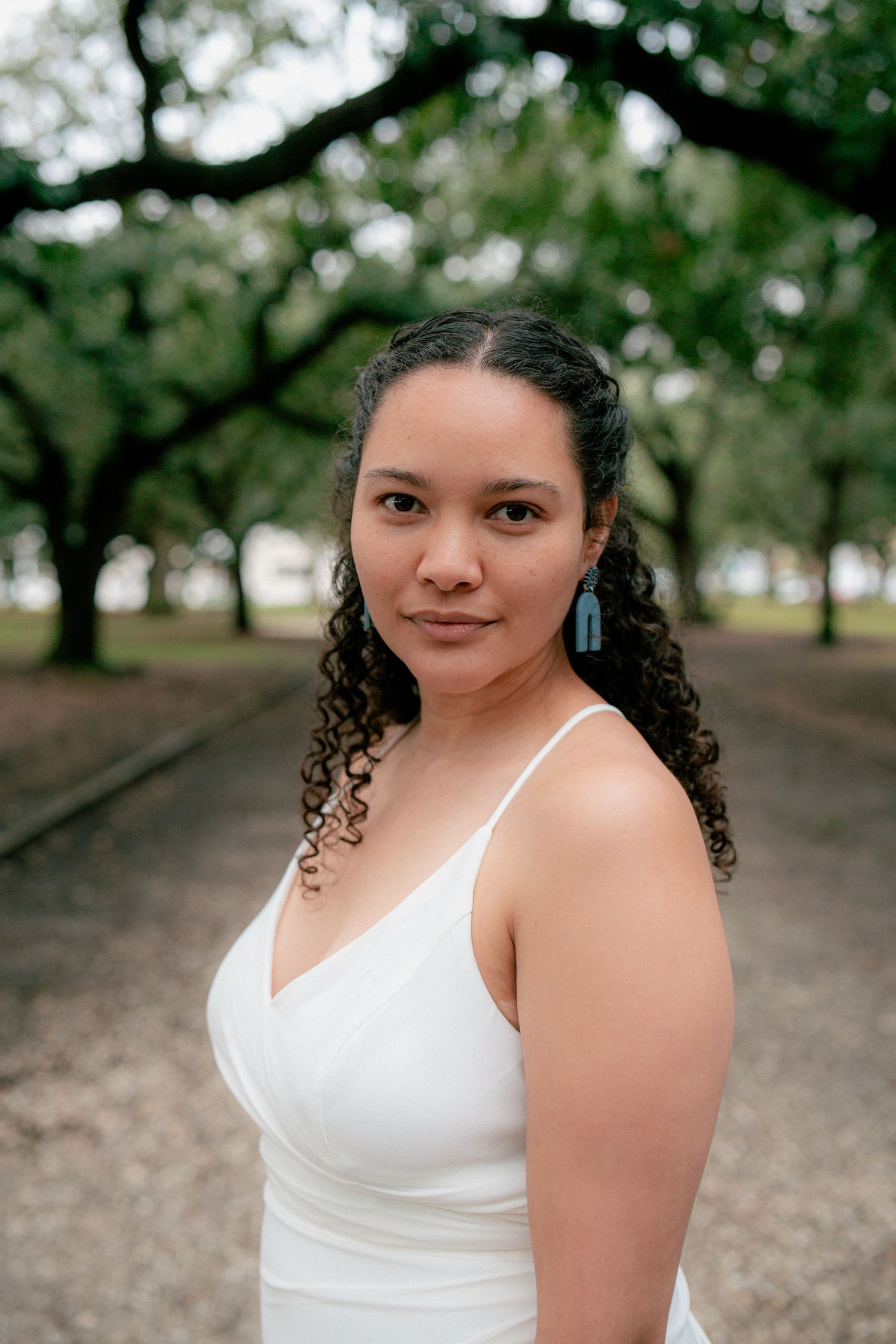 Portrait of a woman with curly hair wearing a white dress and teal earrings in a tree-lined park.