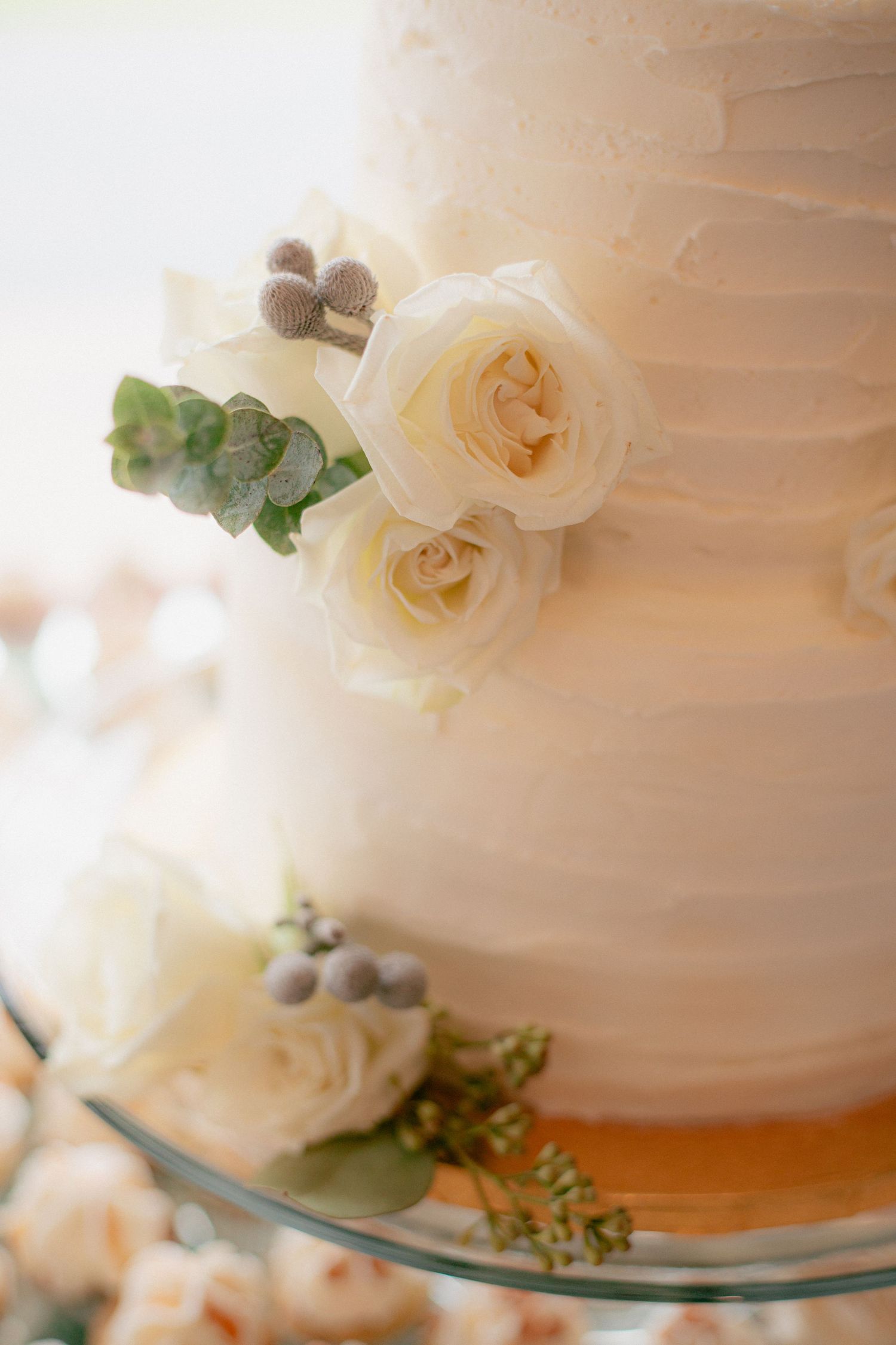 Close-up of a white wedding cake tier decorated with cream roses, greenish leaves, and silver berries.