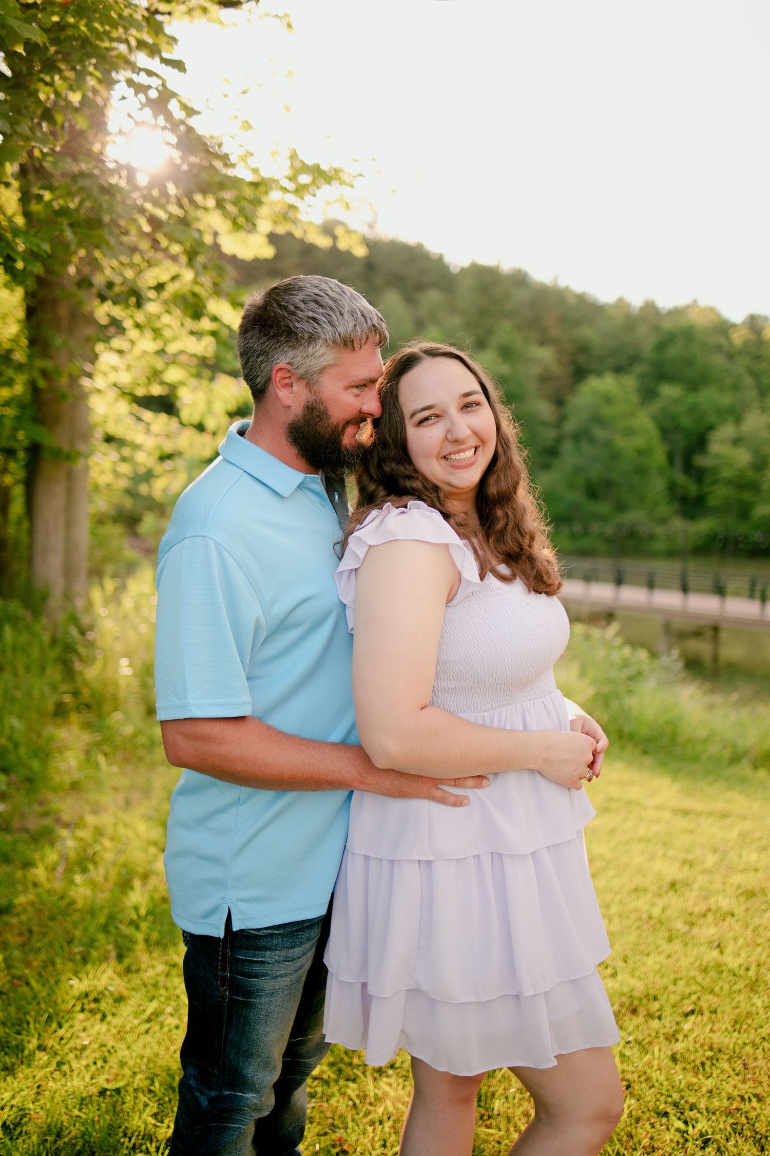A couple embraces outdoors at golden hour, woman in white dress, man in blue shirt, lush green trees in background.