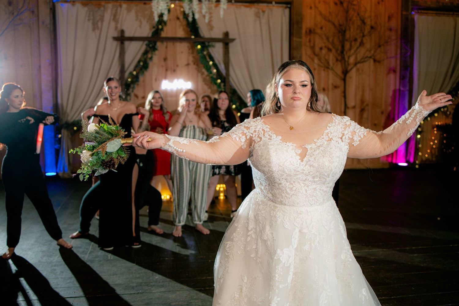 Bride in lace gown tossing bouquet to bridesmaids in rustic barn wedding venue.