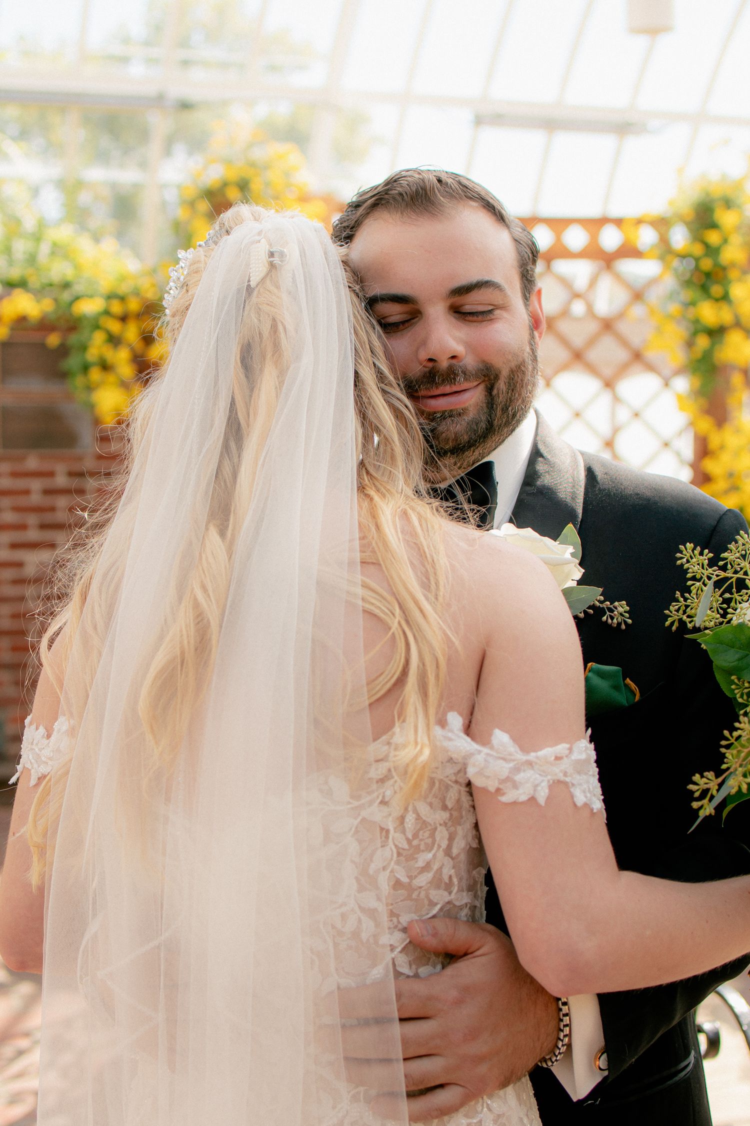 Emotional groom embraces his bride in a greenhouse wedding venue surrounded by vibrant yellow flowers.