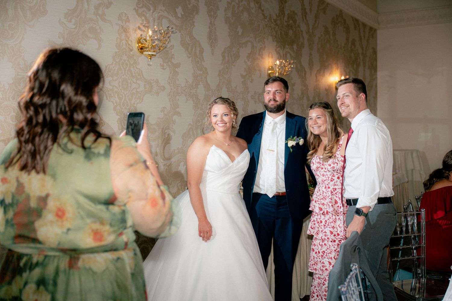 Bride and groom pose for a photo with wedding guests in an elegant wallpapered reception venue.