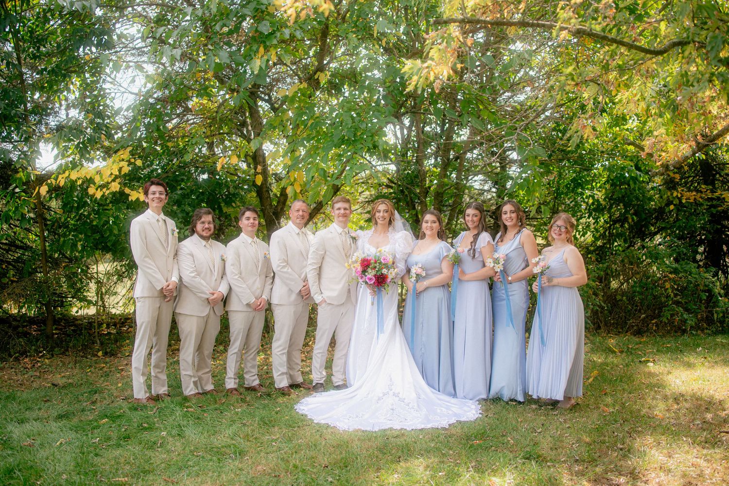 Wedding party posing outdoors, bridesmaids in light blue dresses and groomsmen in tan suits.