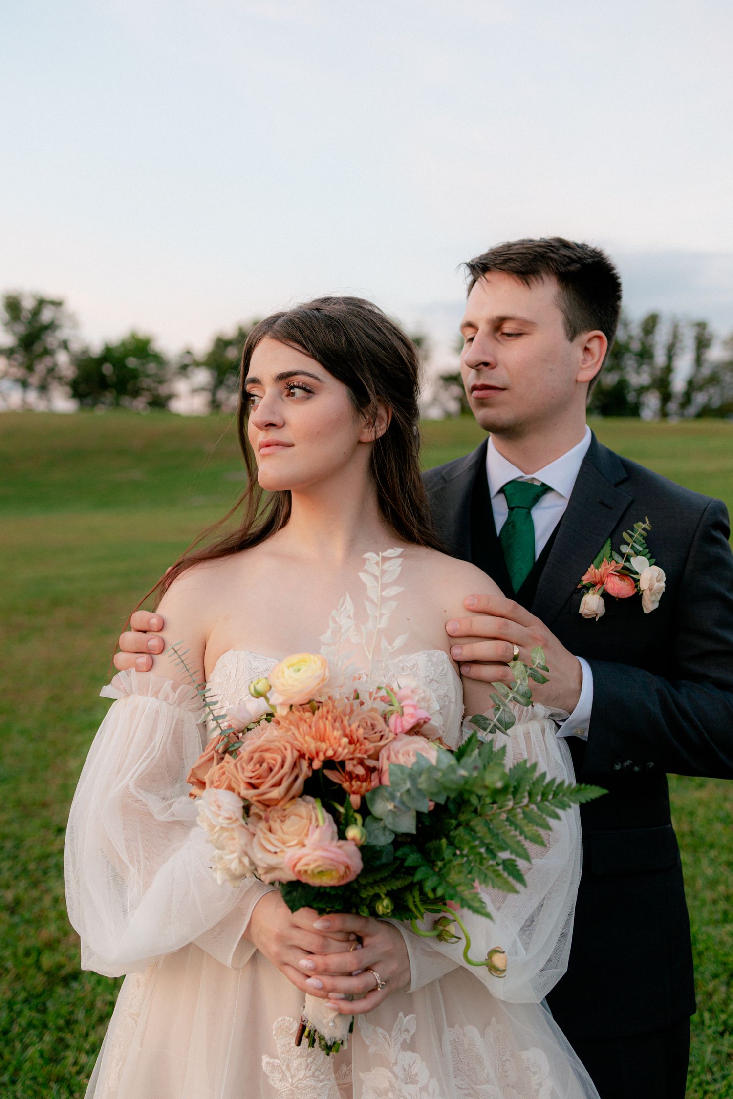 Bride in sheer white gown holds peach bouquet as groom in dark suit with green tie embraces her shoulders outdoors.
