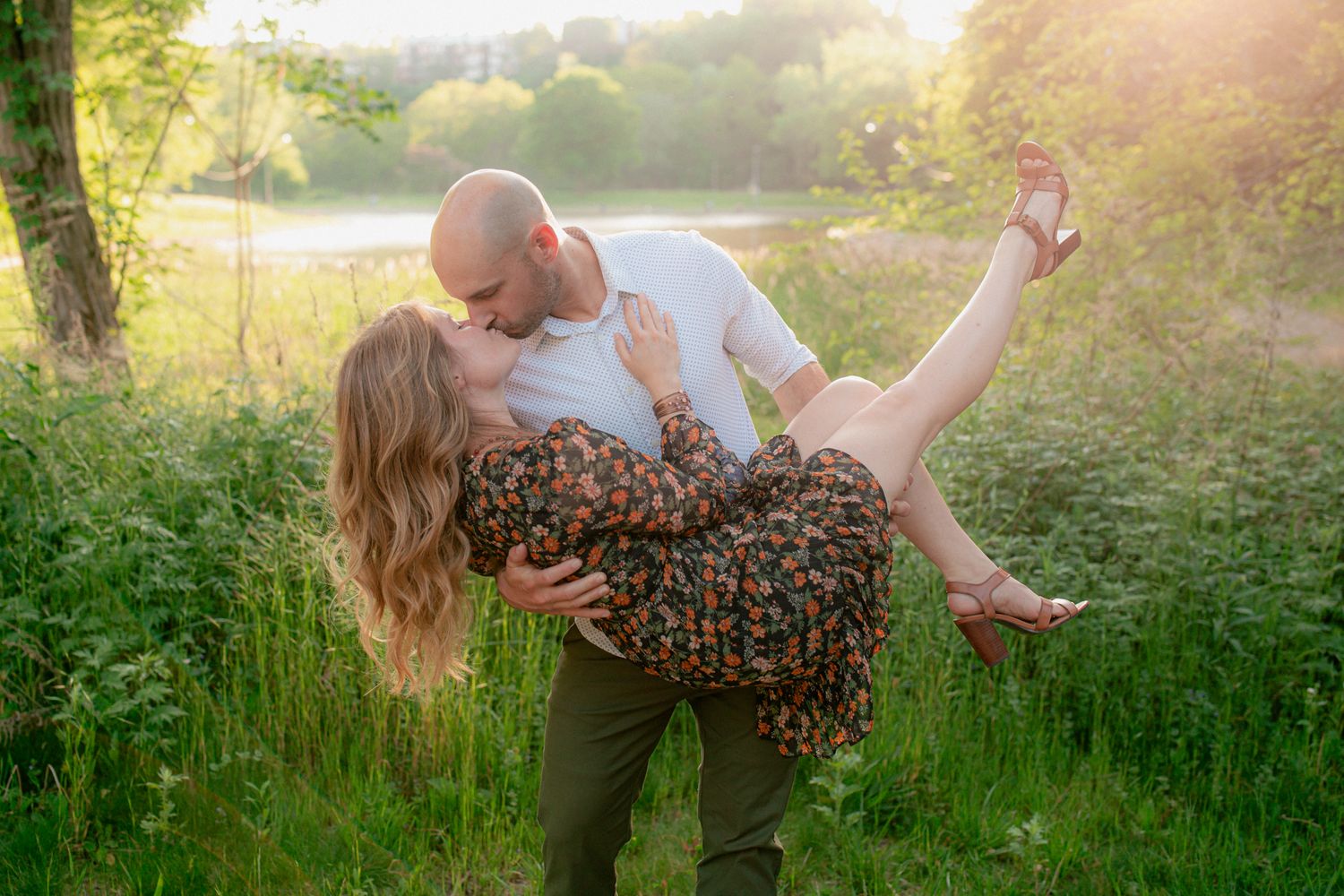 Couple sharing a romantic kiss outdoors, man lifting woman in floral dress in lush green meadow at golden hour.