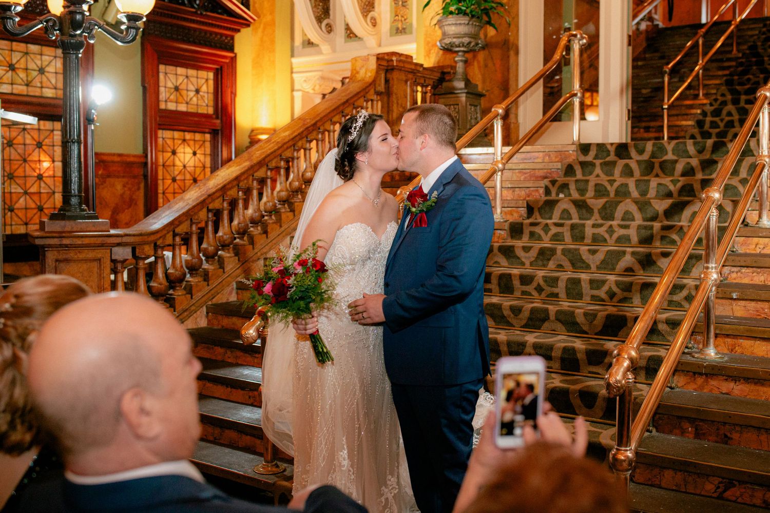Newlyweds share first kiss on ornate staircase as wedding guests capture the romantic moment.