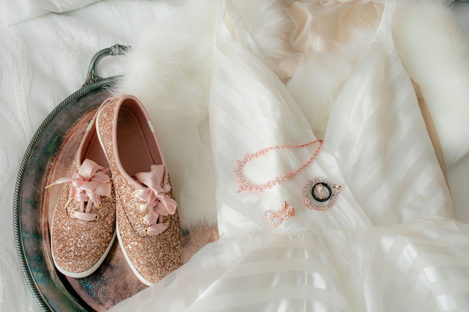 Rose gold glitter wedding shoes, necklace, and rings on white bridal dress.