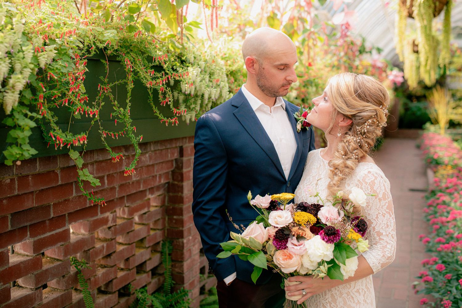 Bride and groom share a tender moment beside a brick wall with lush greenery.